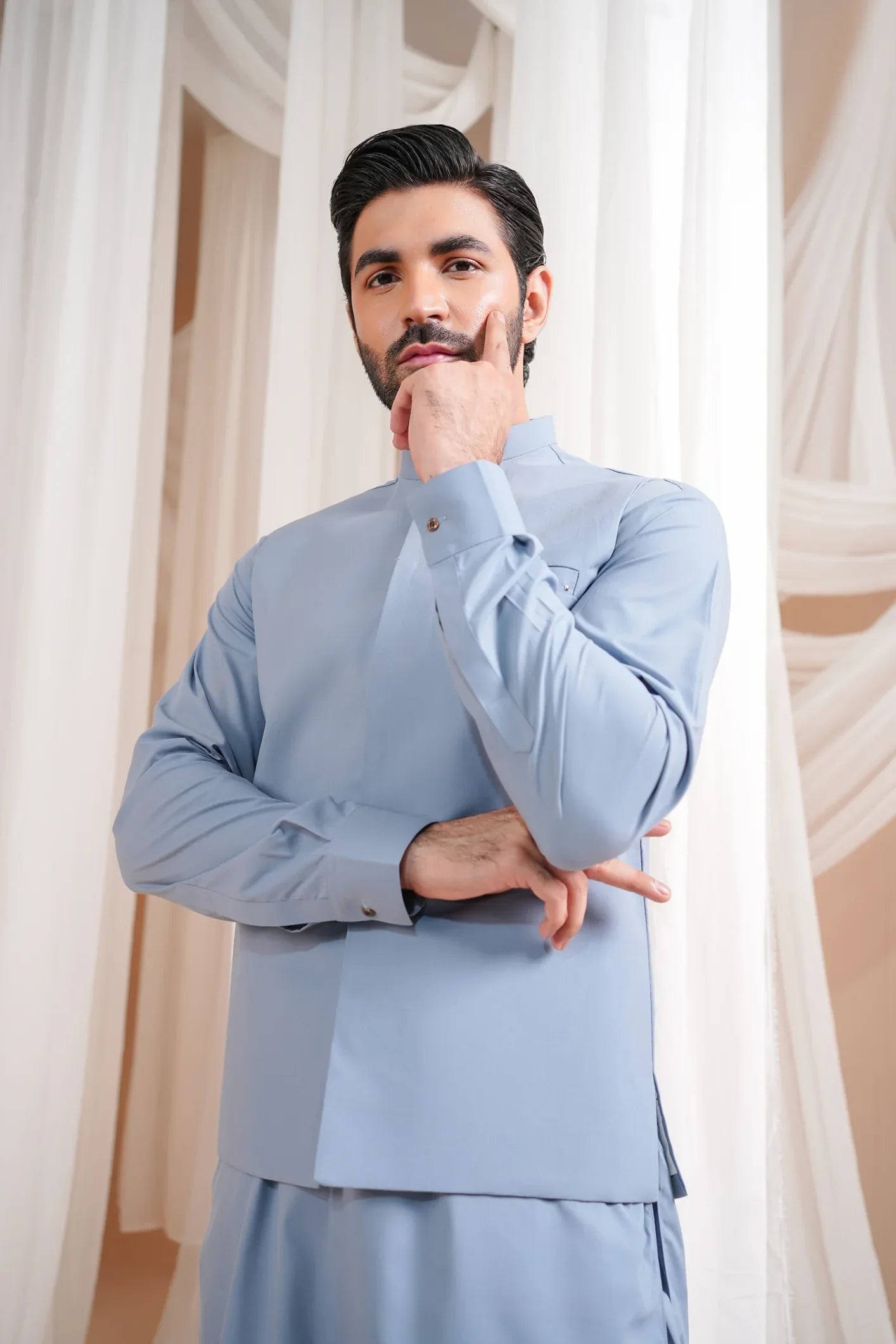 A man wearing a straight cut waistcoat in a sapphire blue color with a mandarin collar and full sleeves, standing in a room with white drapes.