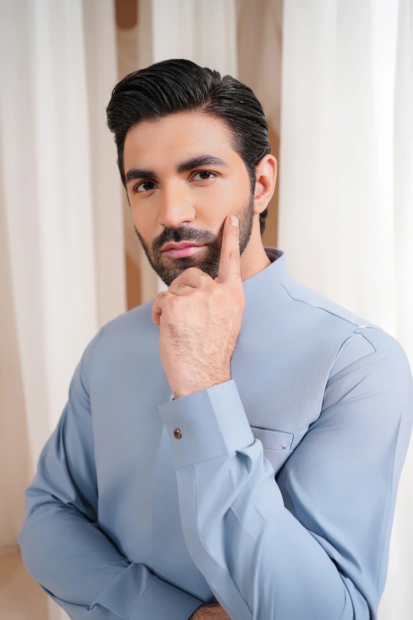 A man wearing a burgundy luxury formal kameez shalwar with a blended waistcoat and full sleeves. He is standing in a room with light curtains behind him.