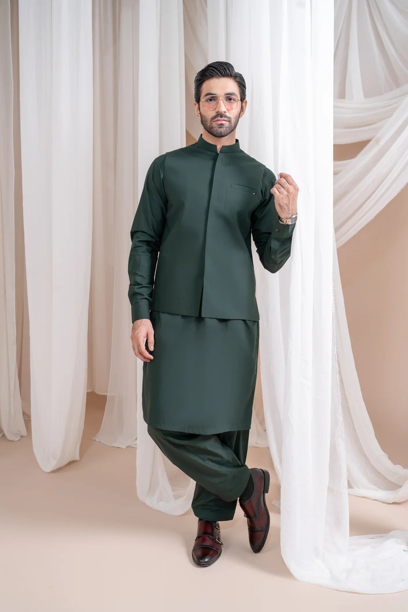 A man wearing a green luxury blended waistcoat with a mandarin collar and straight cut design, standing in a room with white drapes in the background.