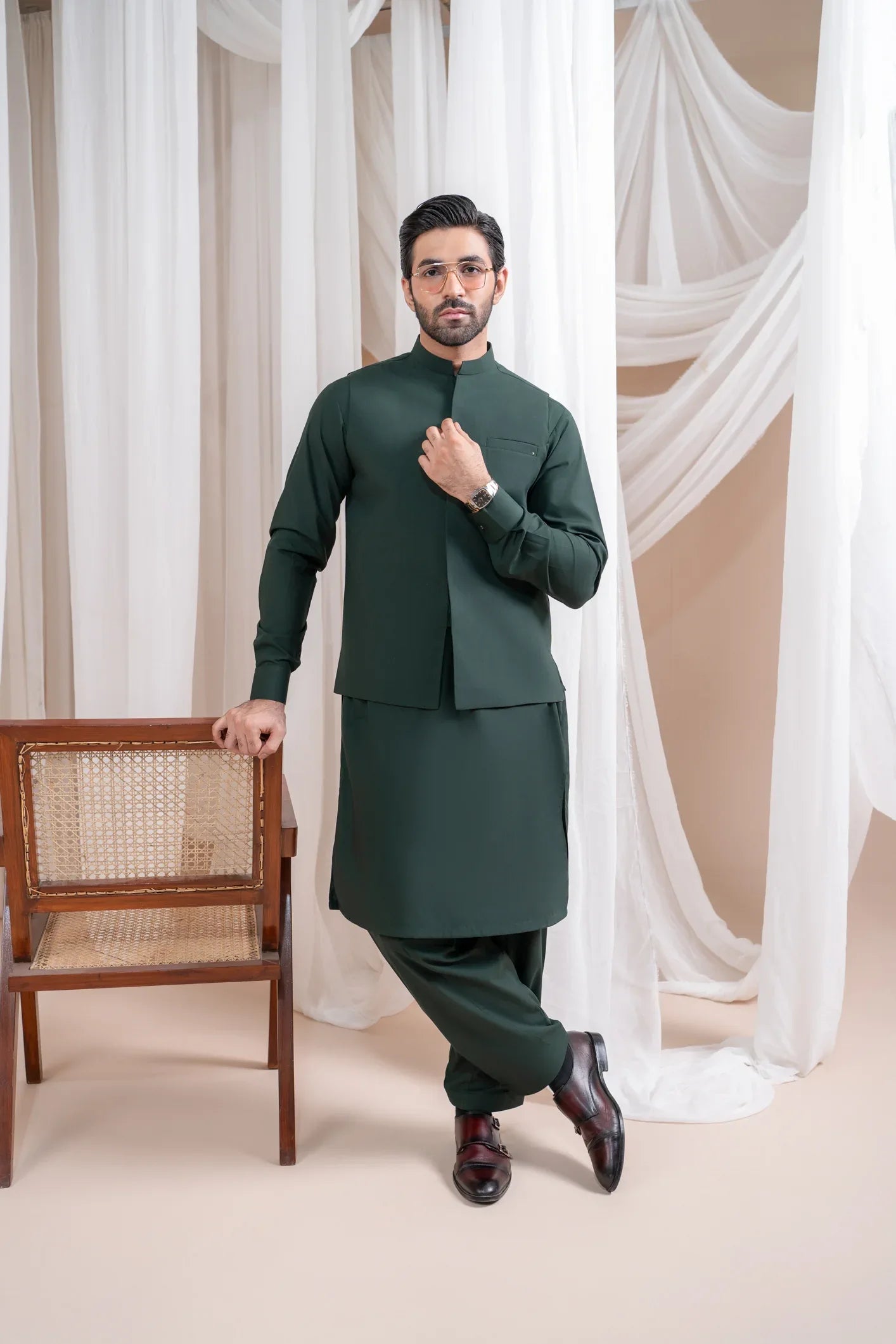 A man wearing a bottle green luxury formal Kameez Shalwar with a patch pocket waistcoat and full sleeves. The outfit is straight cut and suited for all-season wear.