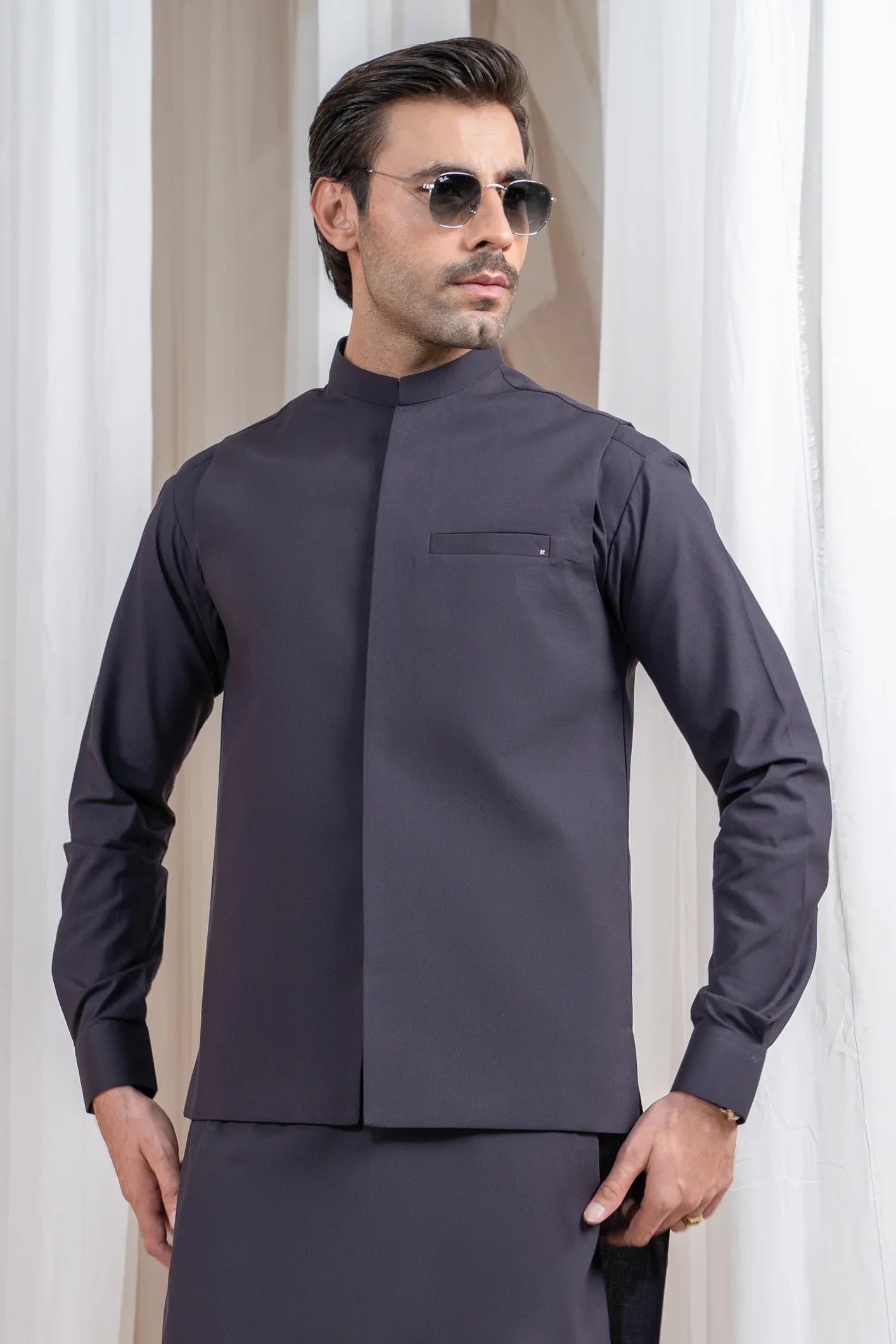 A man wearing a burgundy luxury formal kameez shalwar with a blended waistcoat and full sleeves. He is standing in a room with light curtains behind him.