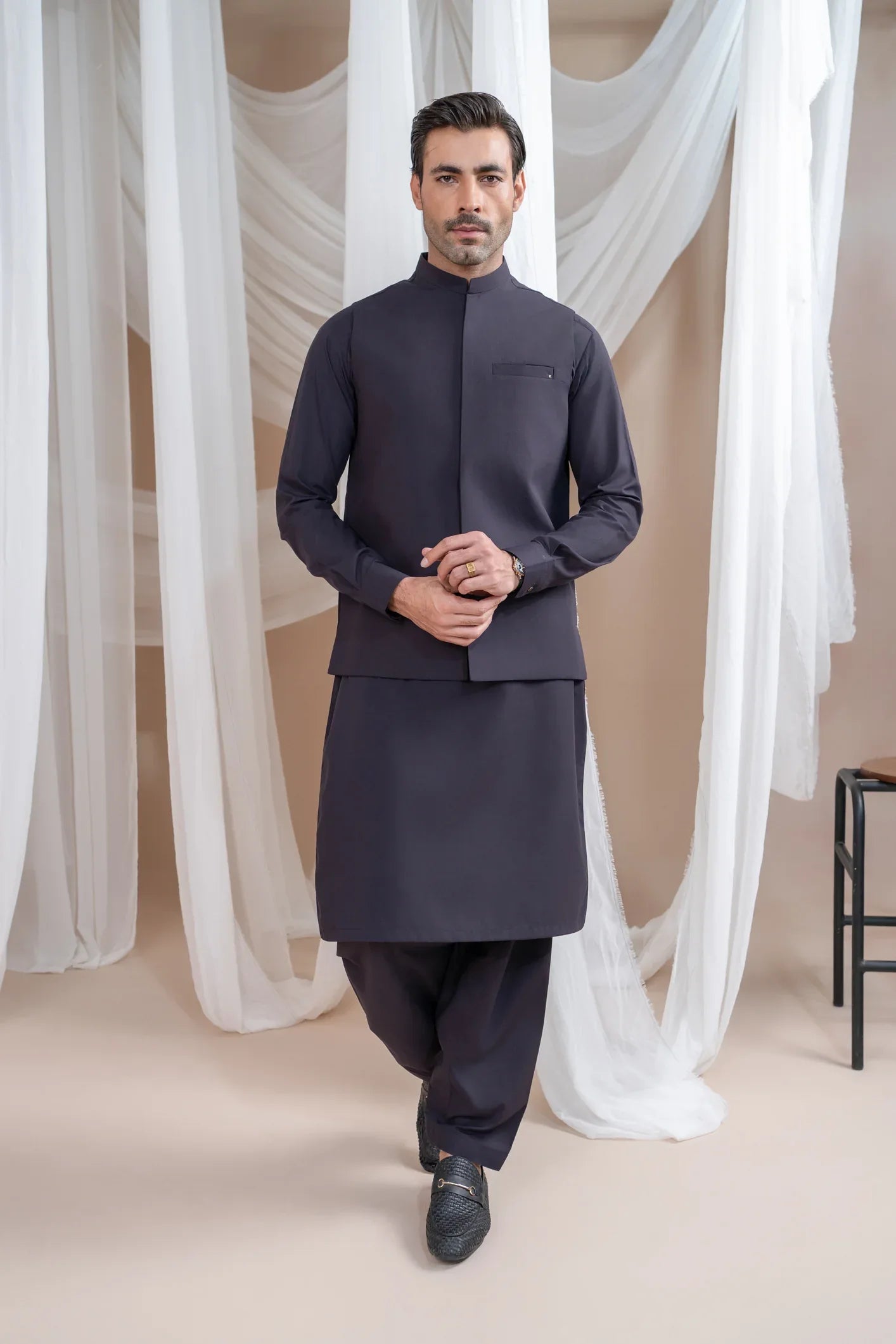 A man wearing a burgundy luxury formal kameez shalwar with a blended waistcoat and full sleeves. He is standing in a room with light curtains behind him.