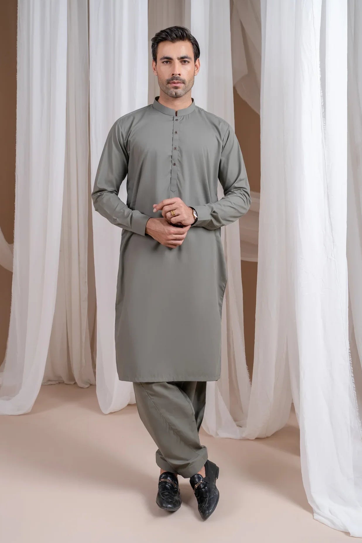 A man standing in a shalwar kameez with a mandarin collar, full sleeves, and a straight kurta and trouser. The background features white drapes and a soft grey background.