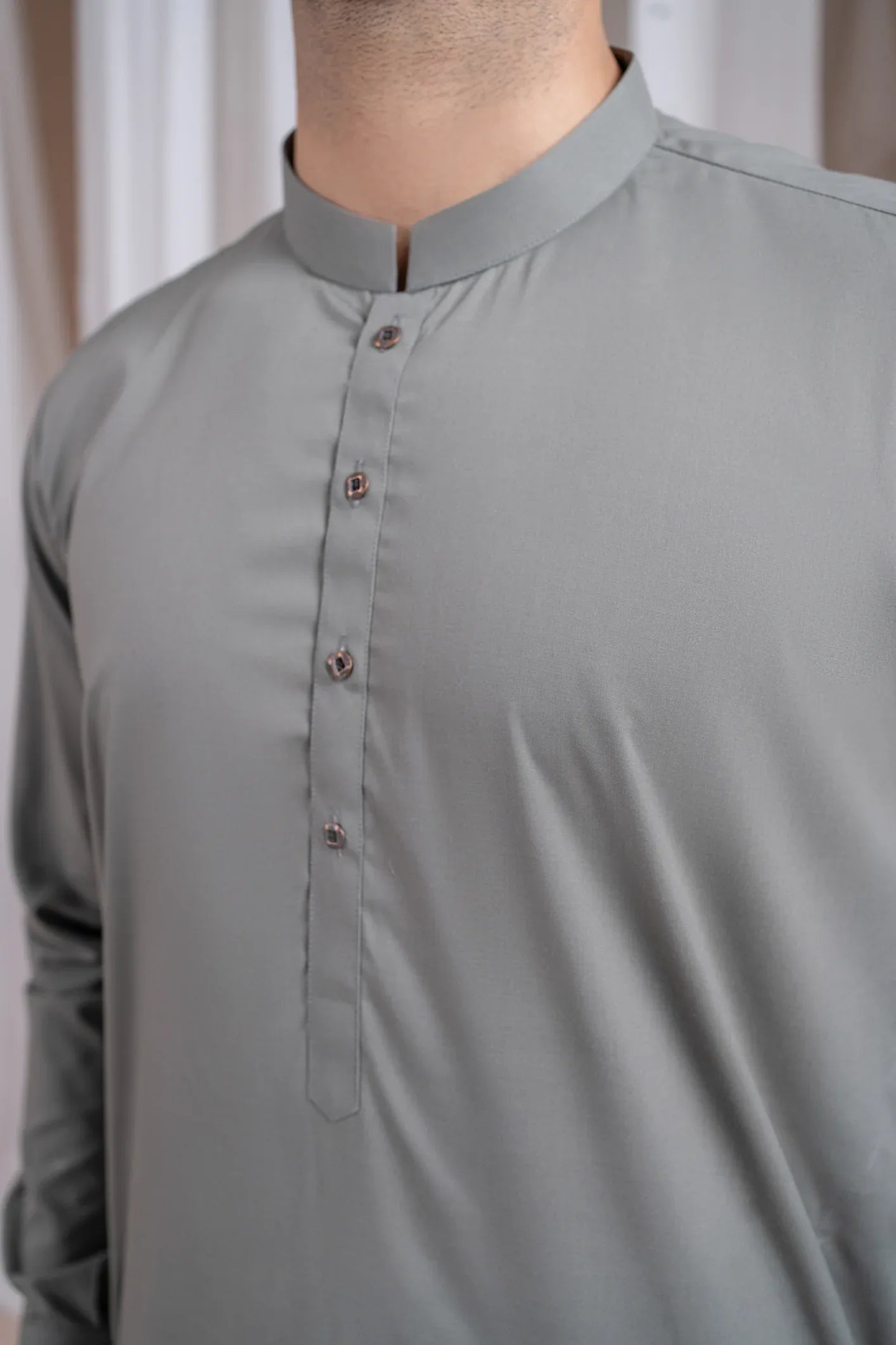 A man standing in a shalwar kameez with a mandarin collar, full sleeves, and a straight kurta and trouser. The background features white drapes and a soft grey background.