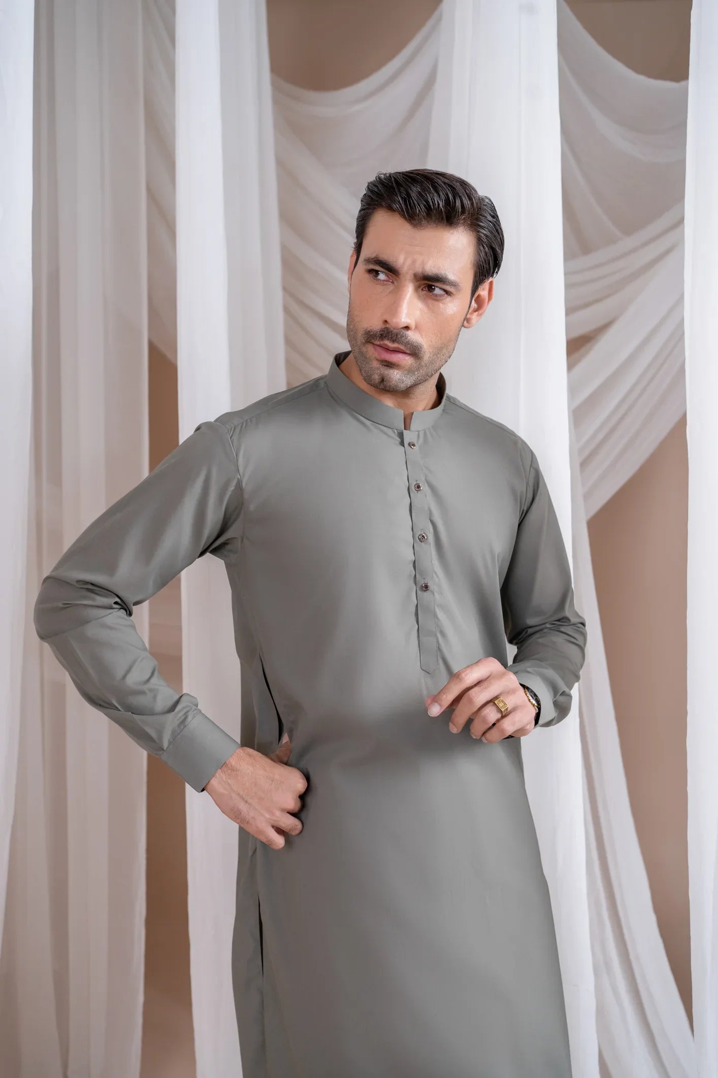A man standing in a shalwar kameez with a mandarin collar, full sleeves, and a straight kurta and trouser. The background features white drapes and a soft grey background.