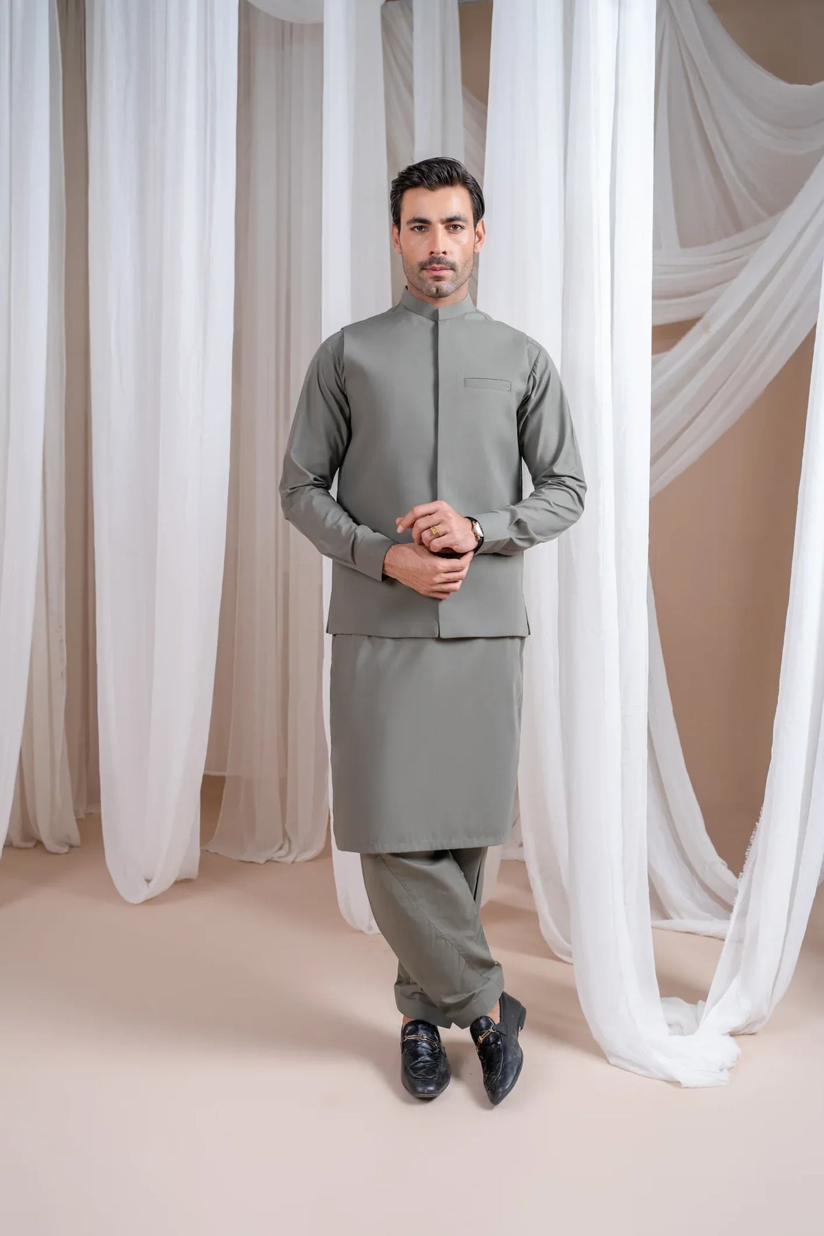 A man wearing a sand grey luxury blended waistcoat with a mandarin collar and full sleeves, paired with matching trousers, standing in a room with white drapes in the background.