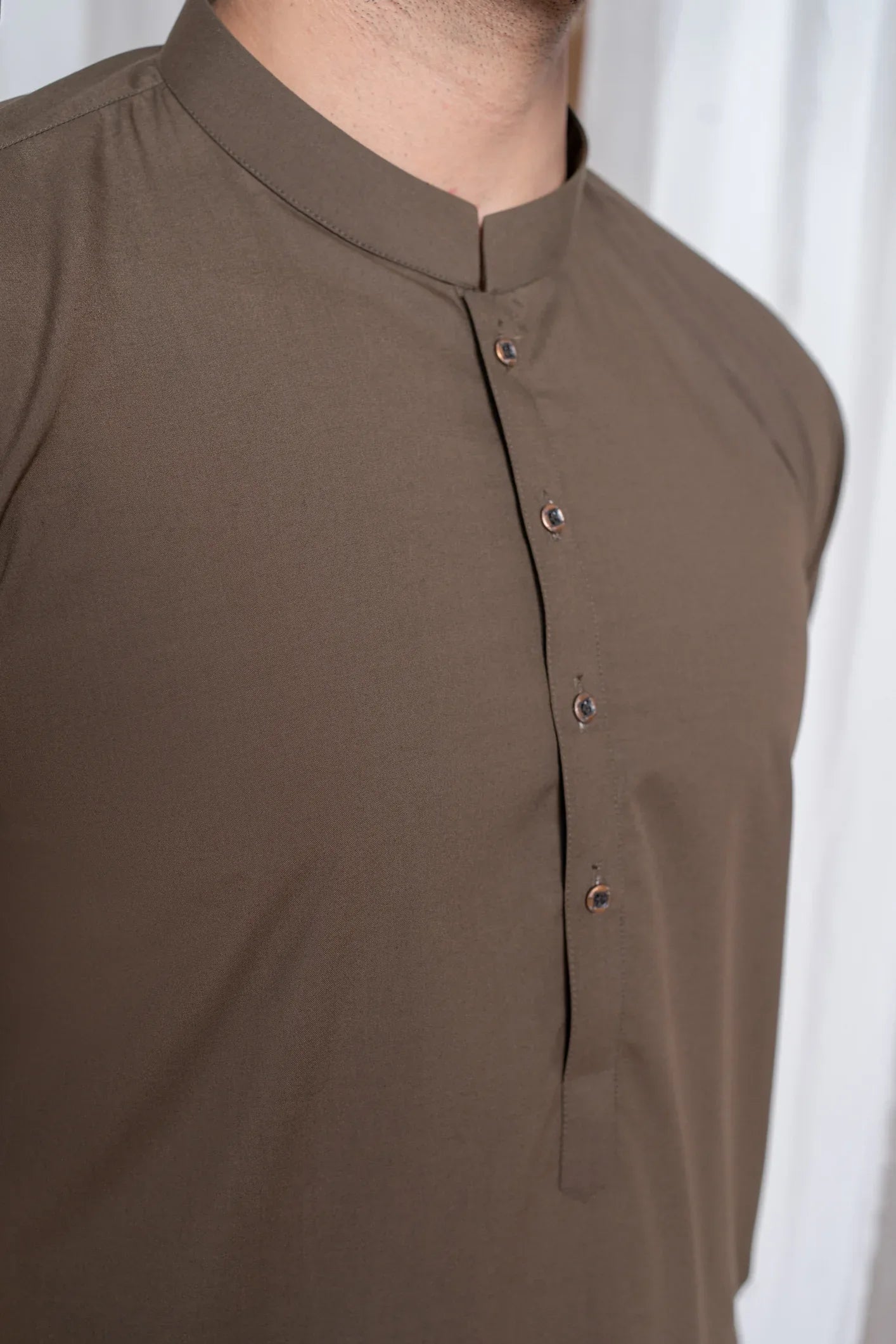 A man wearing a light brown luxury blended kurta shalwar set with a mandarin collar, full sleeves, and a straight kurta and trouser design, standing in a room with white draped backgrounds.