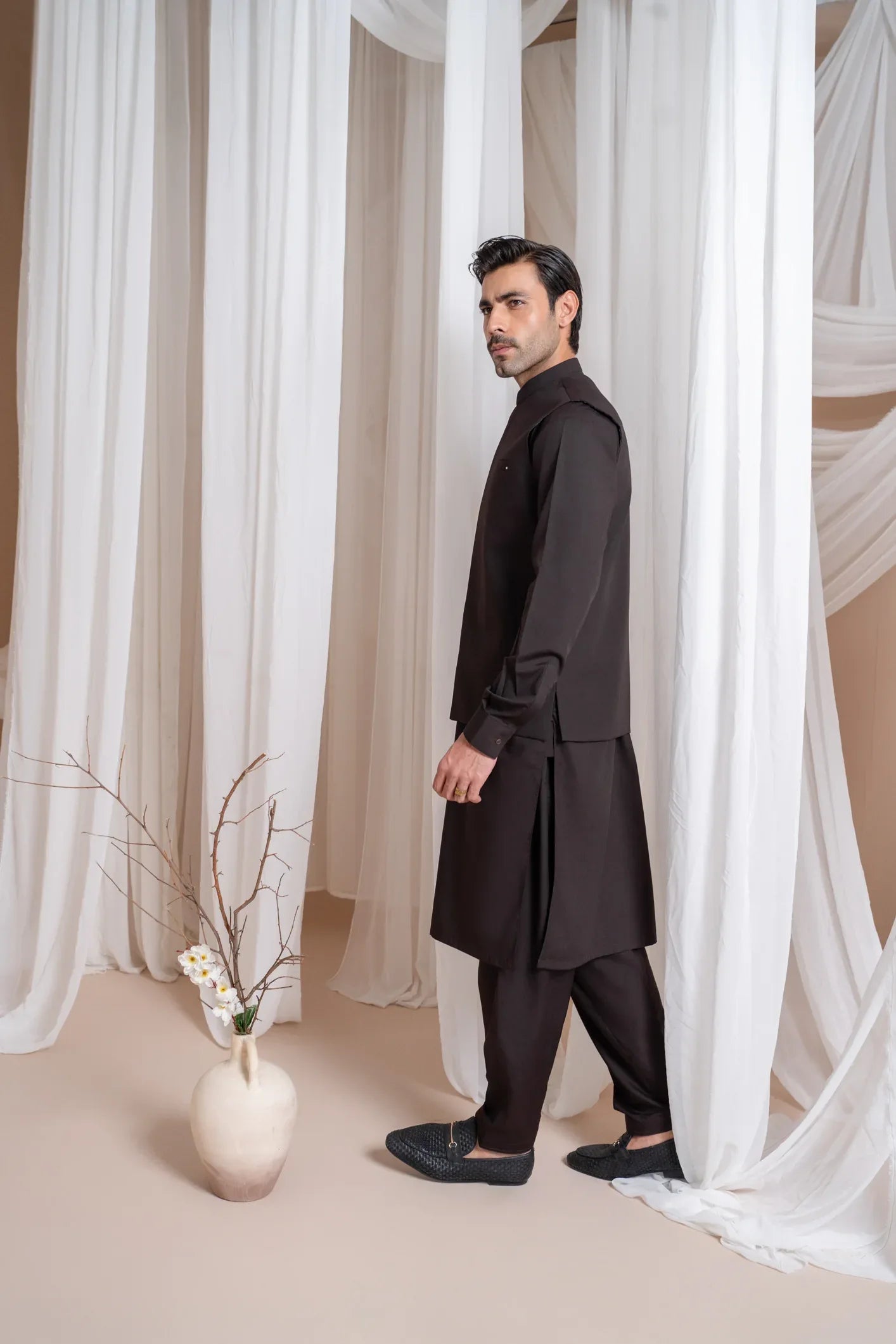 A man wearing a dark brown straight cut kameez shalwar with a blended waistcoat standing indoors with curtains in the background.