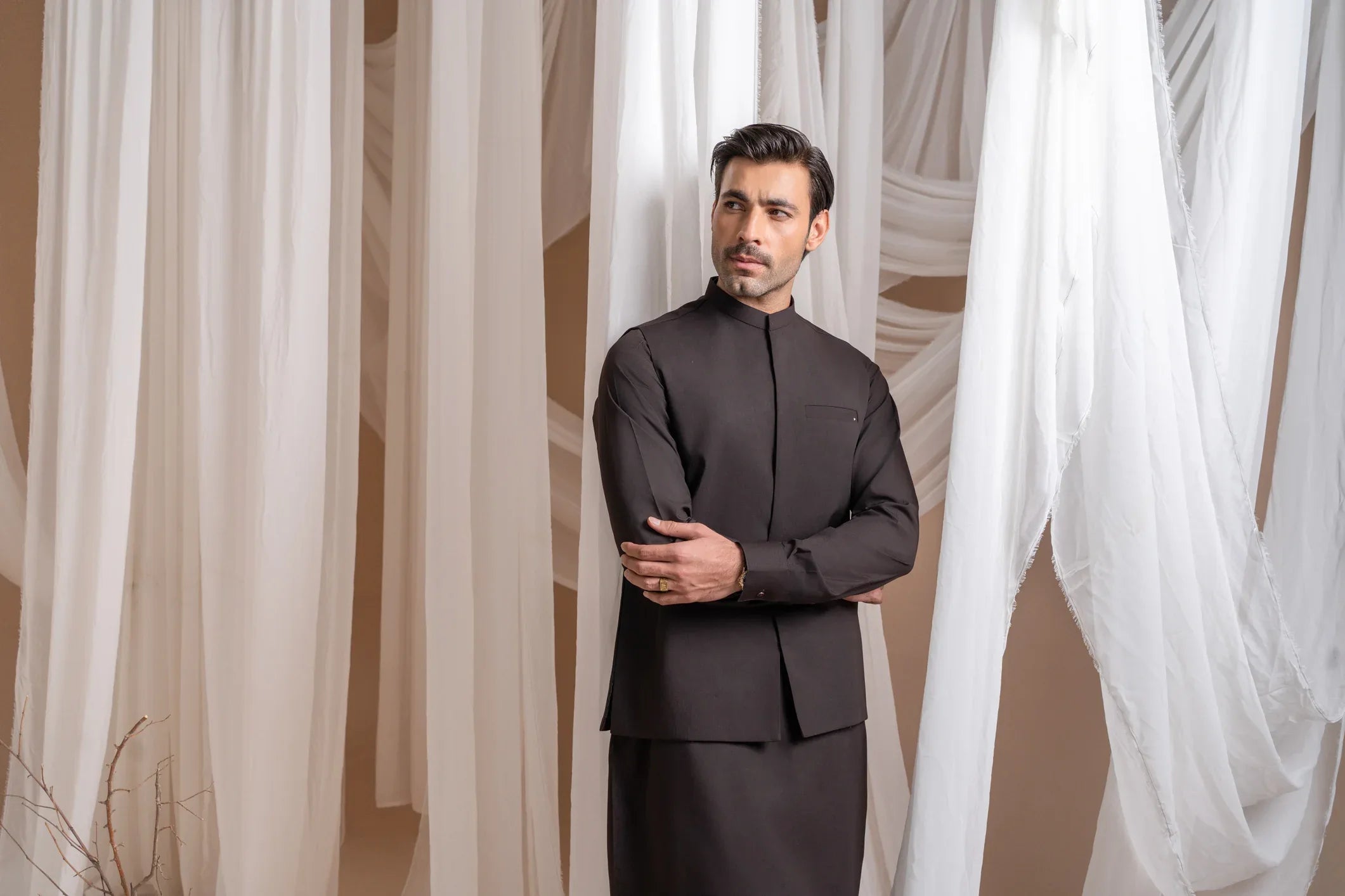 A man wearing a dark brown straight cut kameez shalwar with a blended waistcoat standing indoors with curtains in the background.