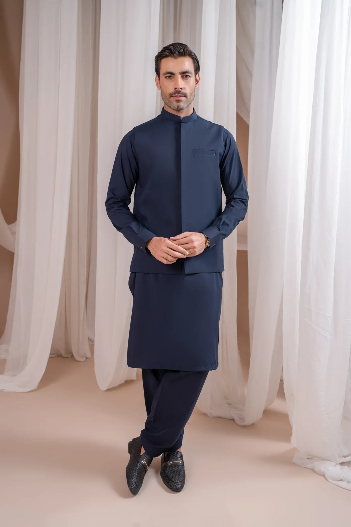 A man wearing a navy blue straight cut waistcoat with a mandarin collar, paired with matching shalwar, standing in a room with white drapes.