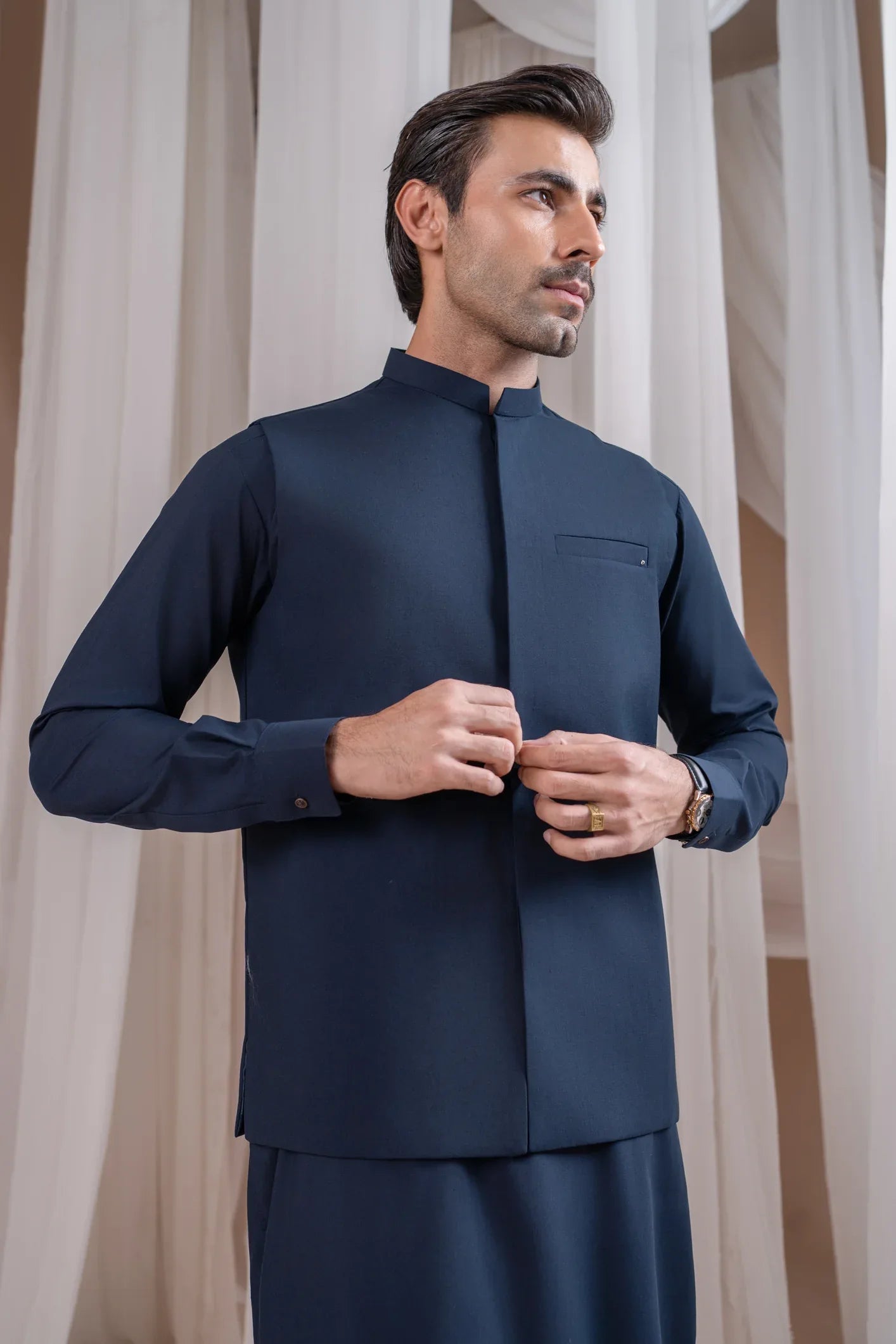 A man wearing a navy blue formal kameez shalwar with a blended waistcoat and full sleeves. He is standing against a neutral backdrop.