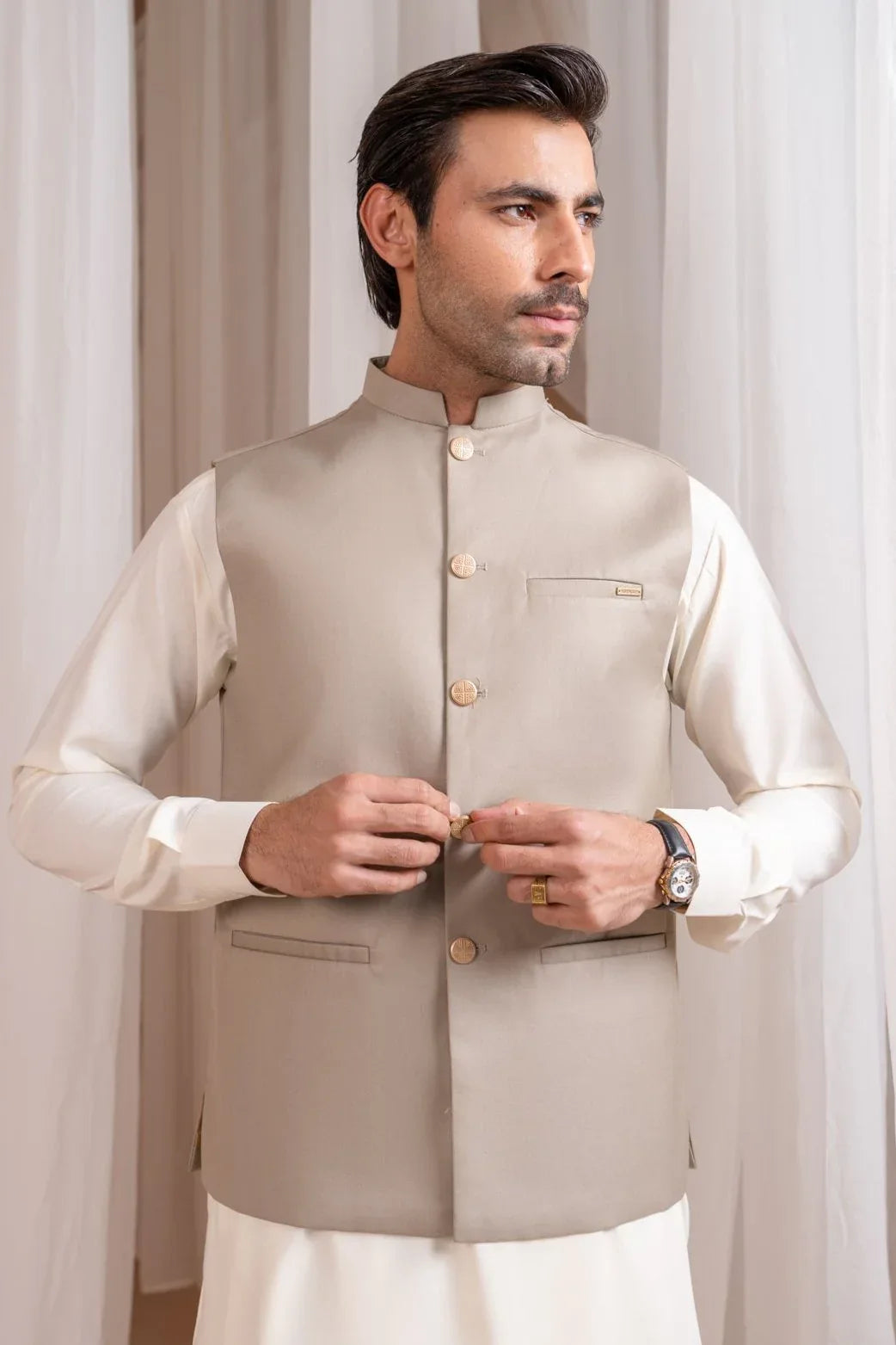 A man wearing a beige suiting waistcoat with button details, standing in a semi-formal setting.