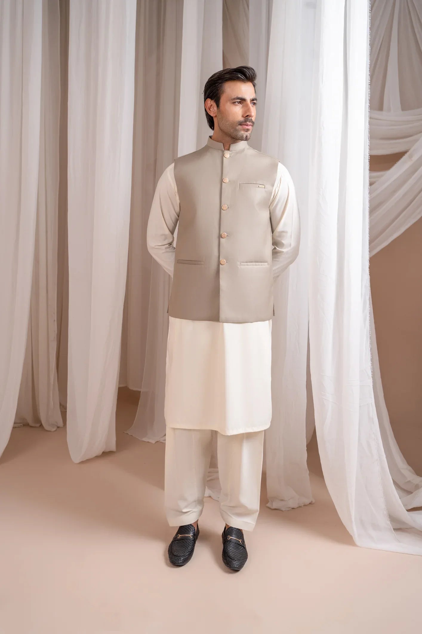 A man wearing a beige suiting waistcoat with button details, standing in a semi-formal setting.