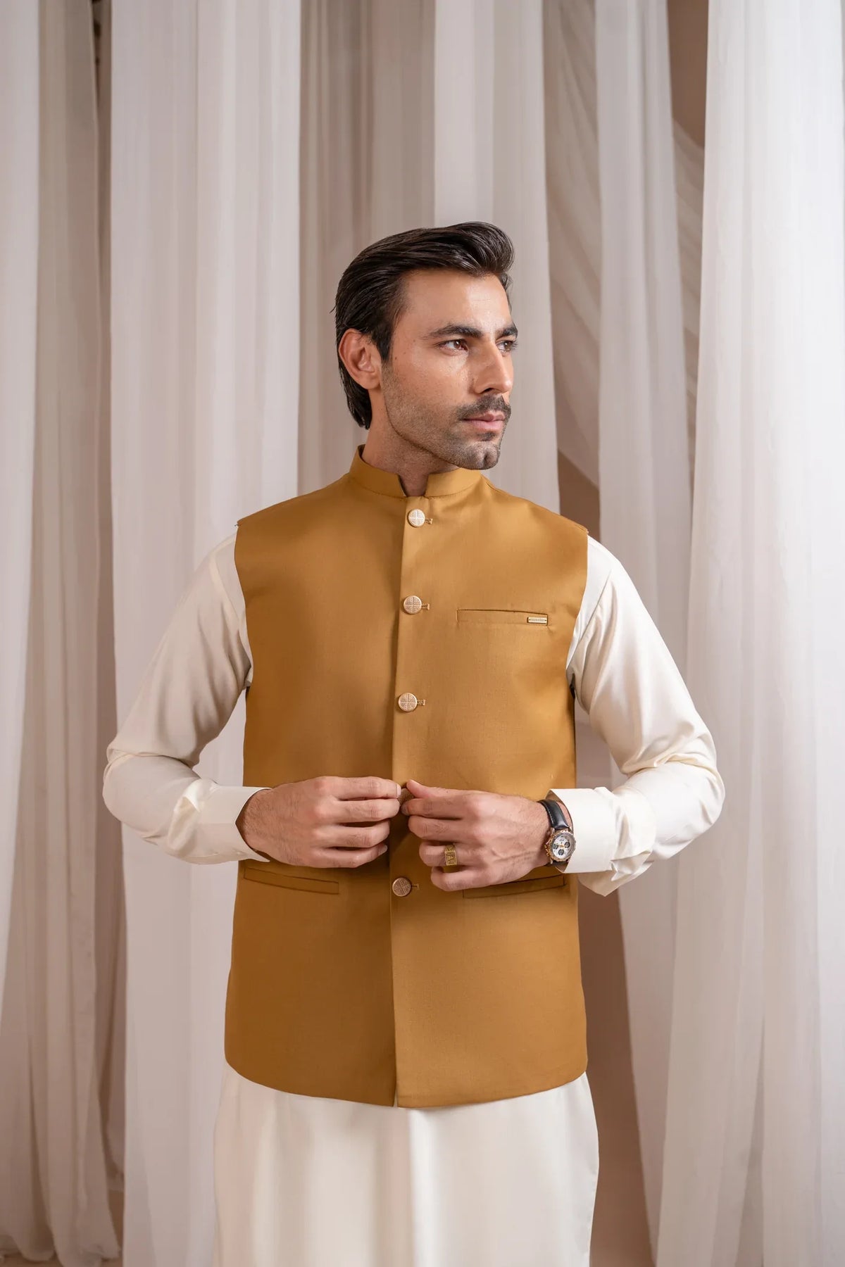 Man wearing a kameez shalwar with a camel brown waistcoat against a white curtain background