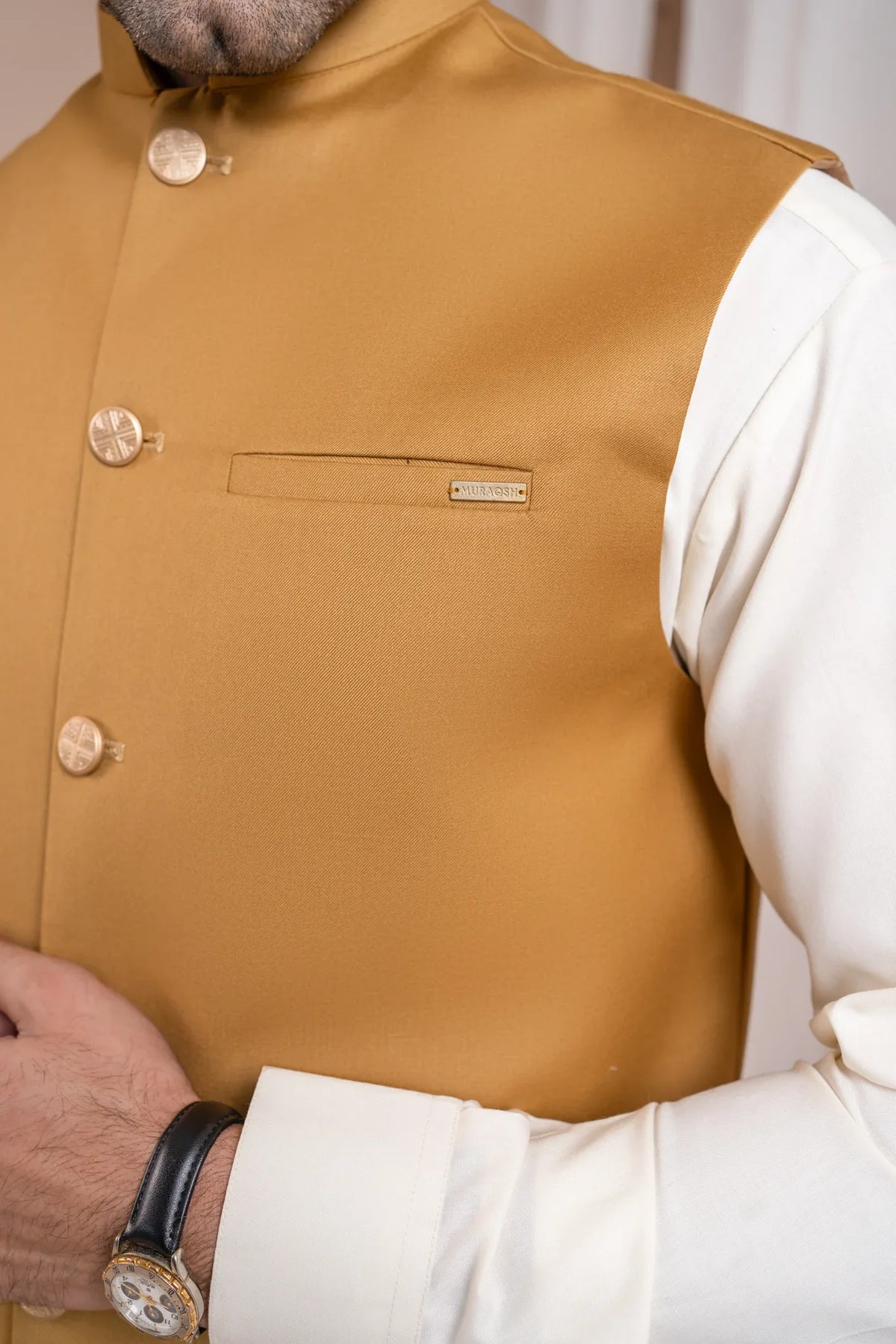 Man wearing a kameez shalwar with a camel brown waistcoat against a white curtain background