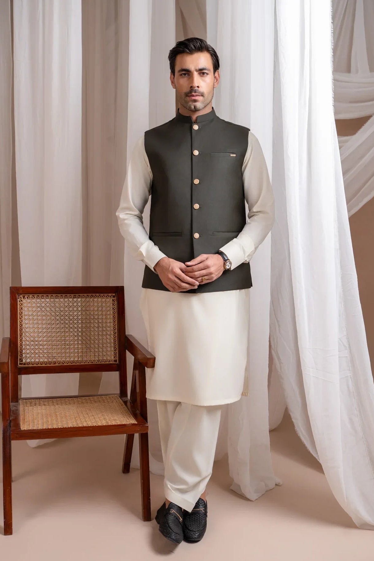 Man wearing a army green suiting waistcoat over bosky white kameez shalwar with a neutral background