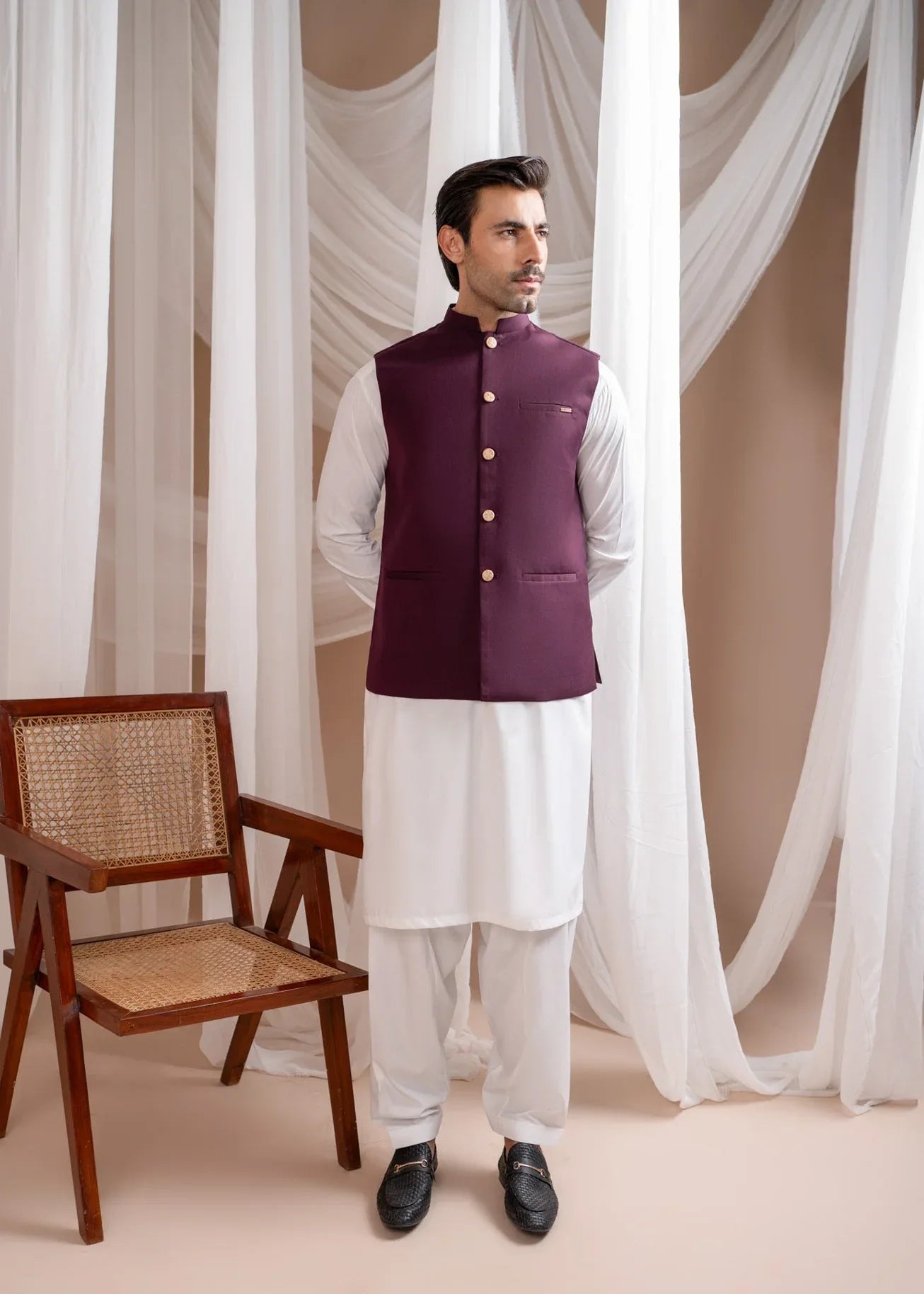Man wearing a burgundy suitinf waistcoat over an off white kameez shalwar, standing in a room with curtains and a chair.