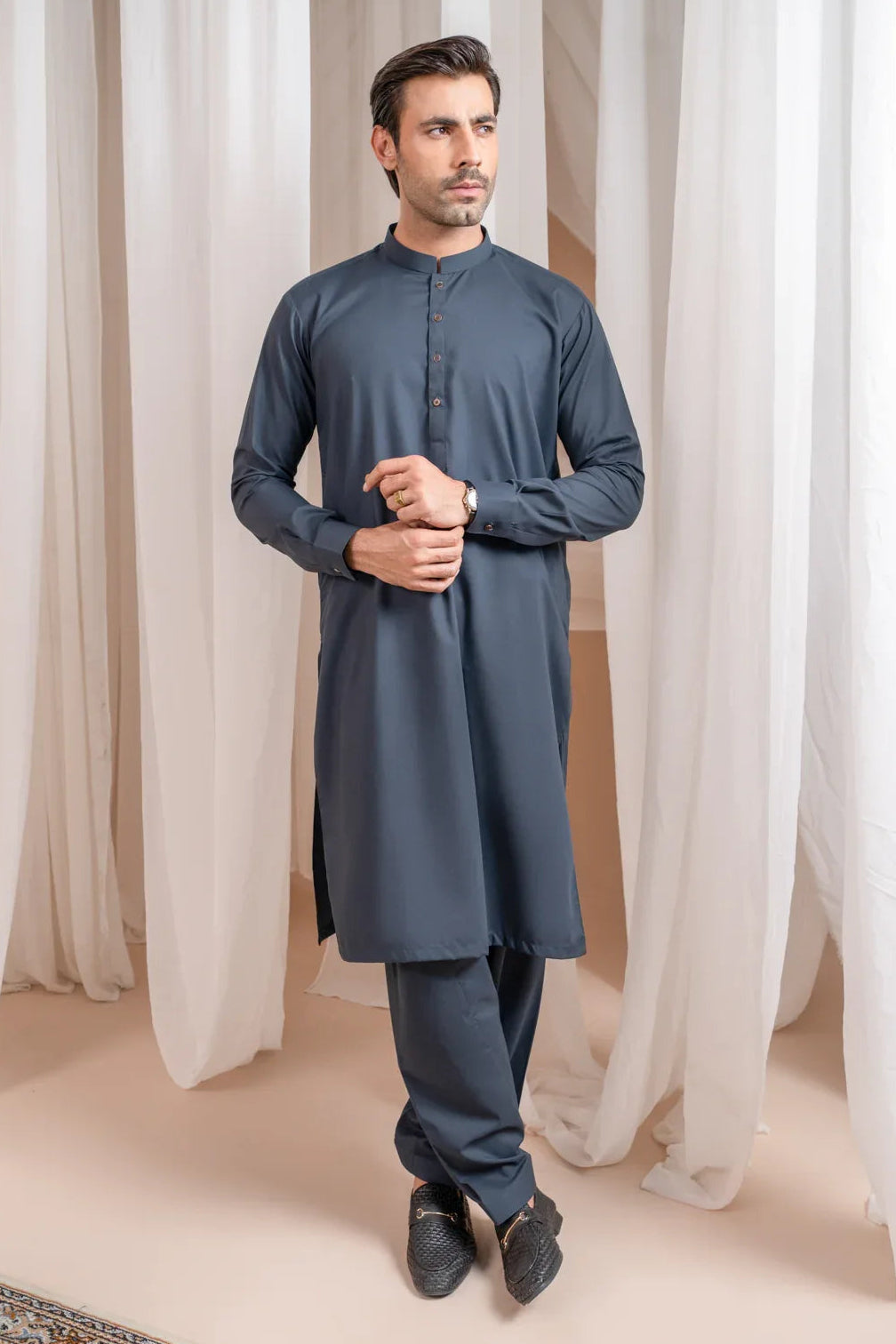 A man standing in a Charcoal grey kameez shalwar with a mandarin collar and full sleeves, in a room with white curtains and a carpeted floor.