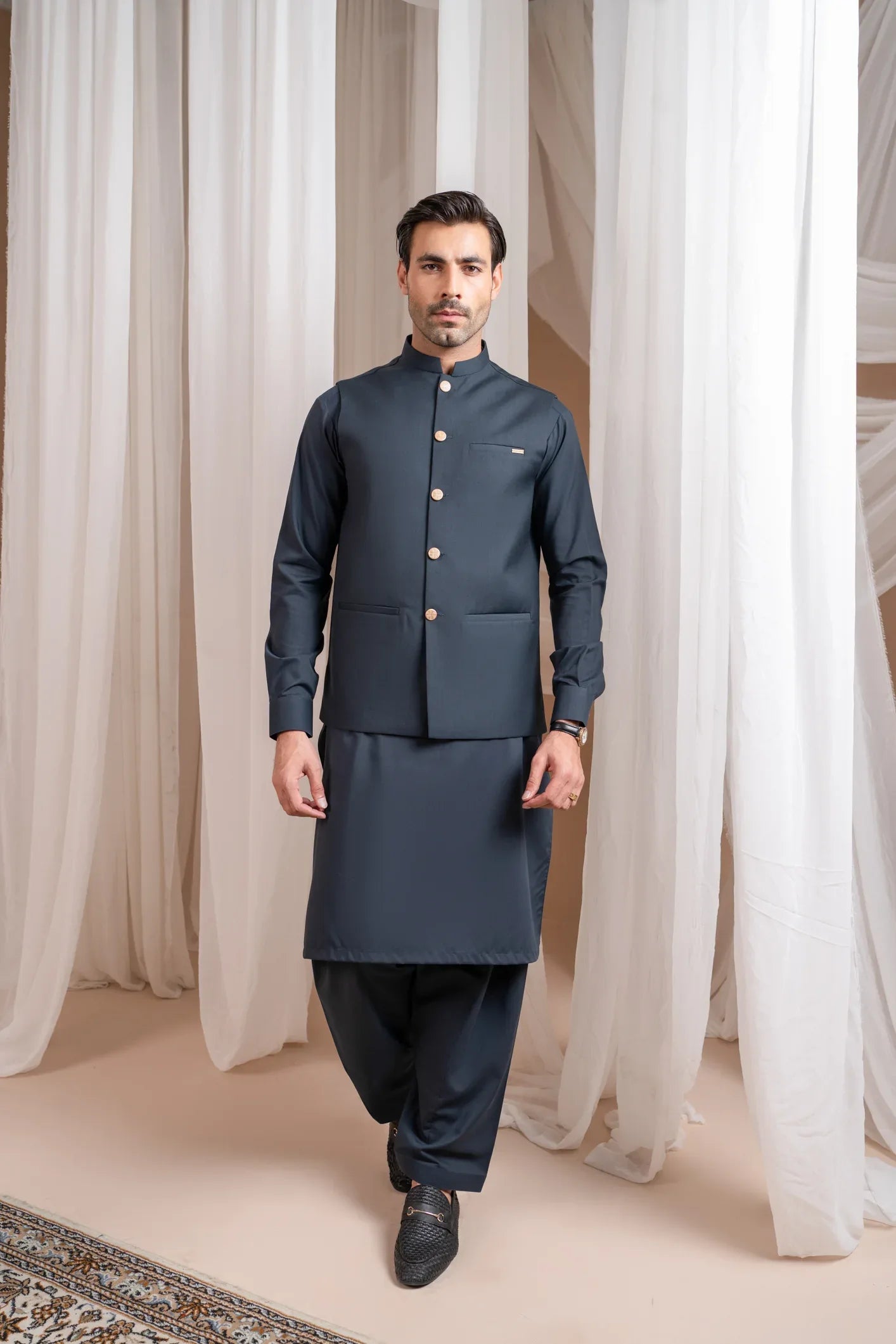 A man standing in a traditional attire, consisting of a charcoal grey kurta shalwar and a matching suiting waistcoat, against a backdrop with white drapes.