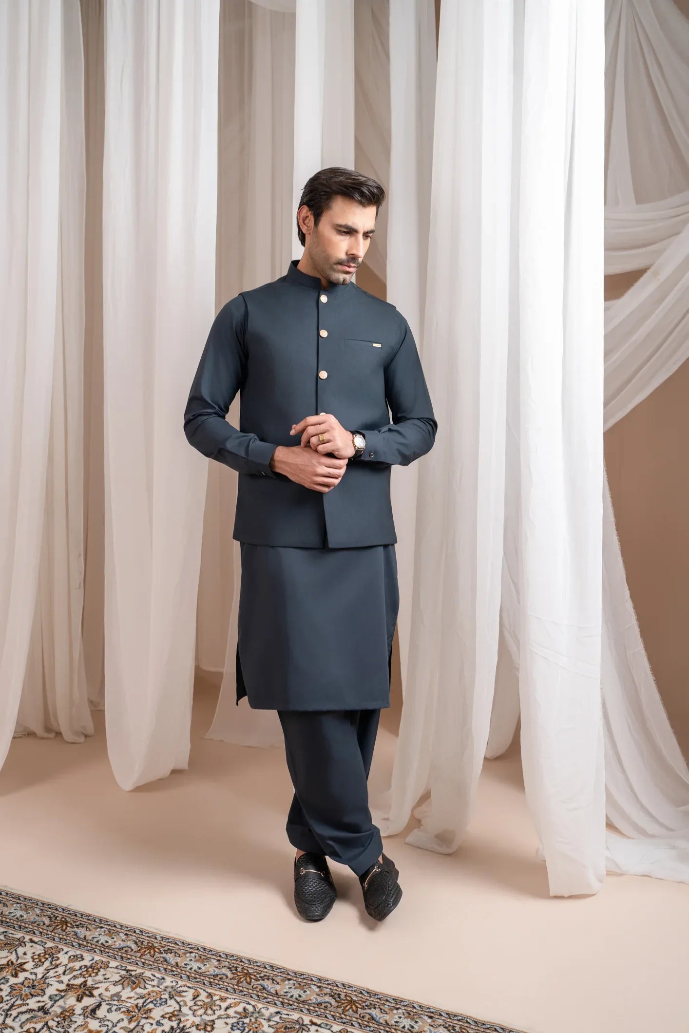A man standing in a traditional attire, consisting of a charcoal grey kurta shalwar and a matching suiting waistcoat, against a backdrop with white drapes.
