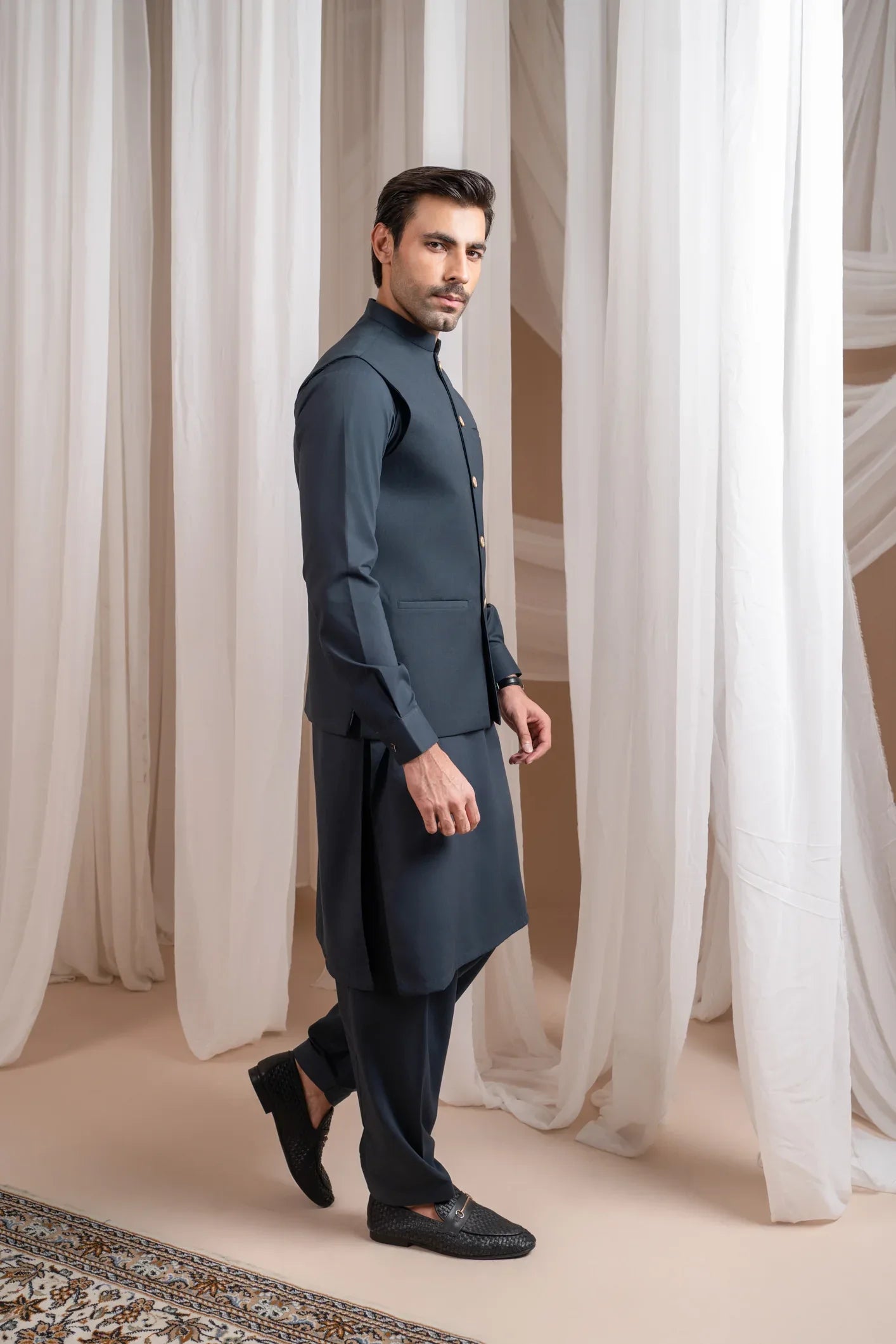 A man standing in a traditional attire, consisting of a charcoal grey kurta shalwar and a matching suiting waistcoat, against a backdrop with white drapes.