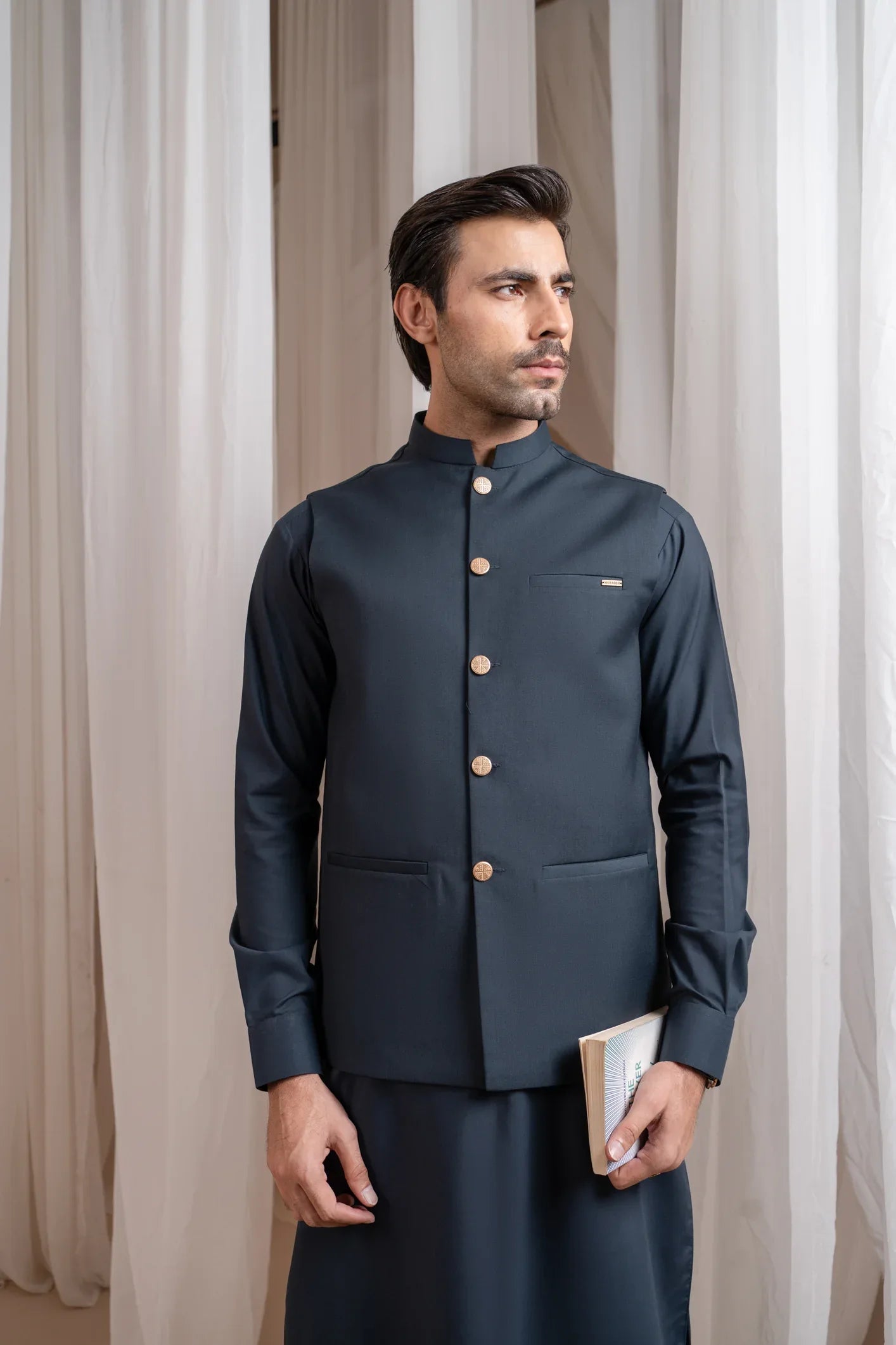 A man standing in a traditional attire, consisting of a charcoal grey kurta shalwar and a matching suiting waistcoat, against a backdrop with white drapes.