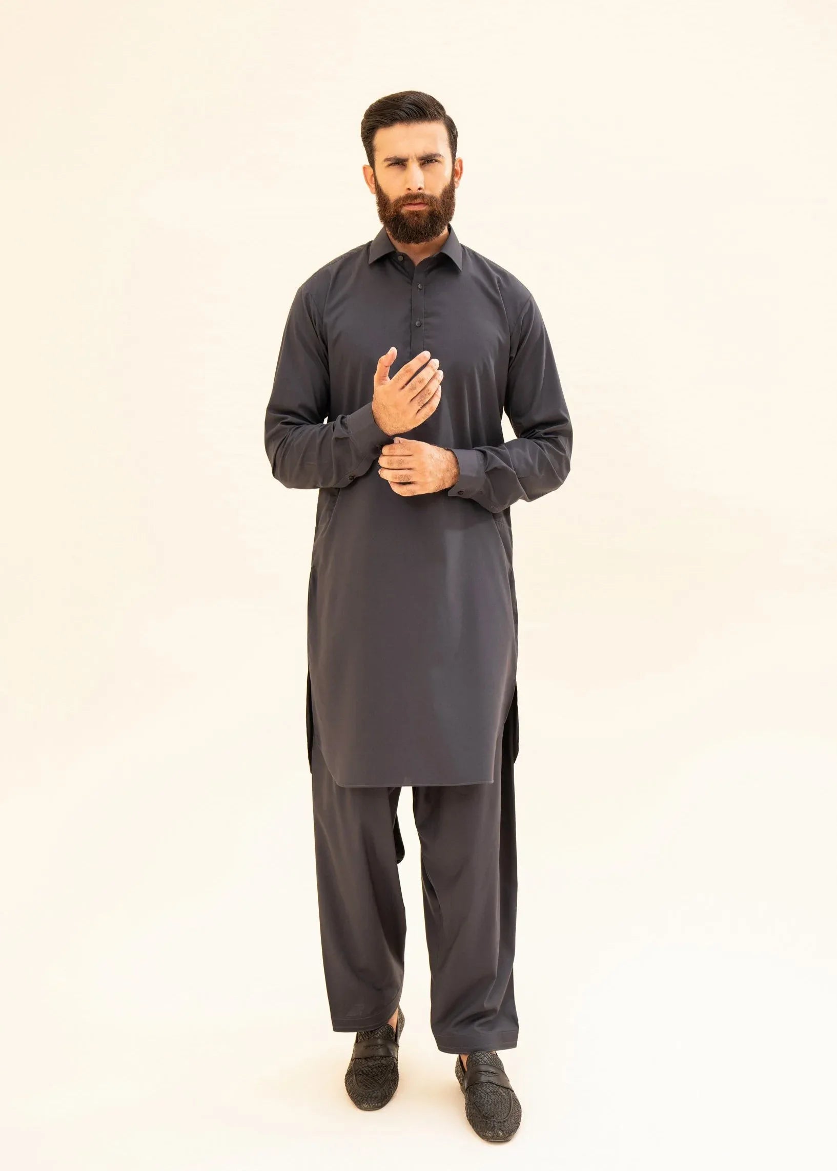 A man wearing a charcoal grey kameez shalwar with a french collar and full sleeves, standing against a white background.