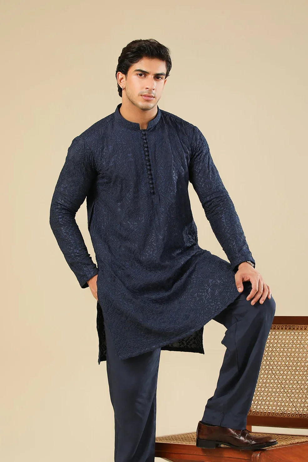Man wearing a navy kurta with pants sitting on a wooden chair against a beige background