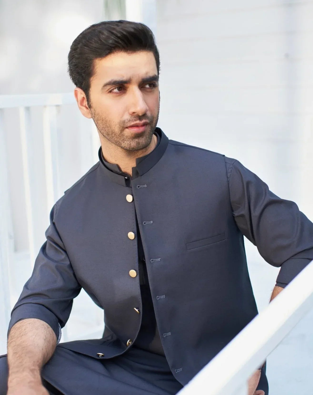 A man wearing a charcoal grey waistcoat with a mandarin collar and a patch pocket, seated with one hand on a white railing, against a background of a white building.