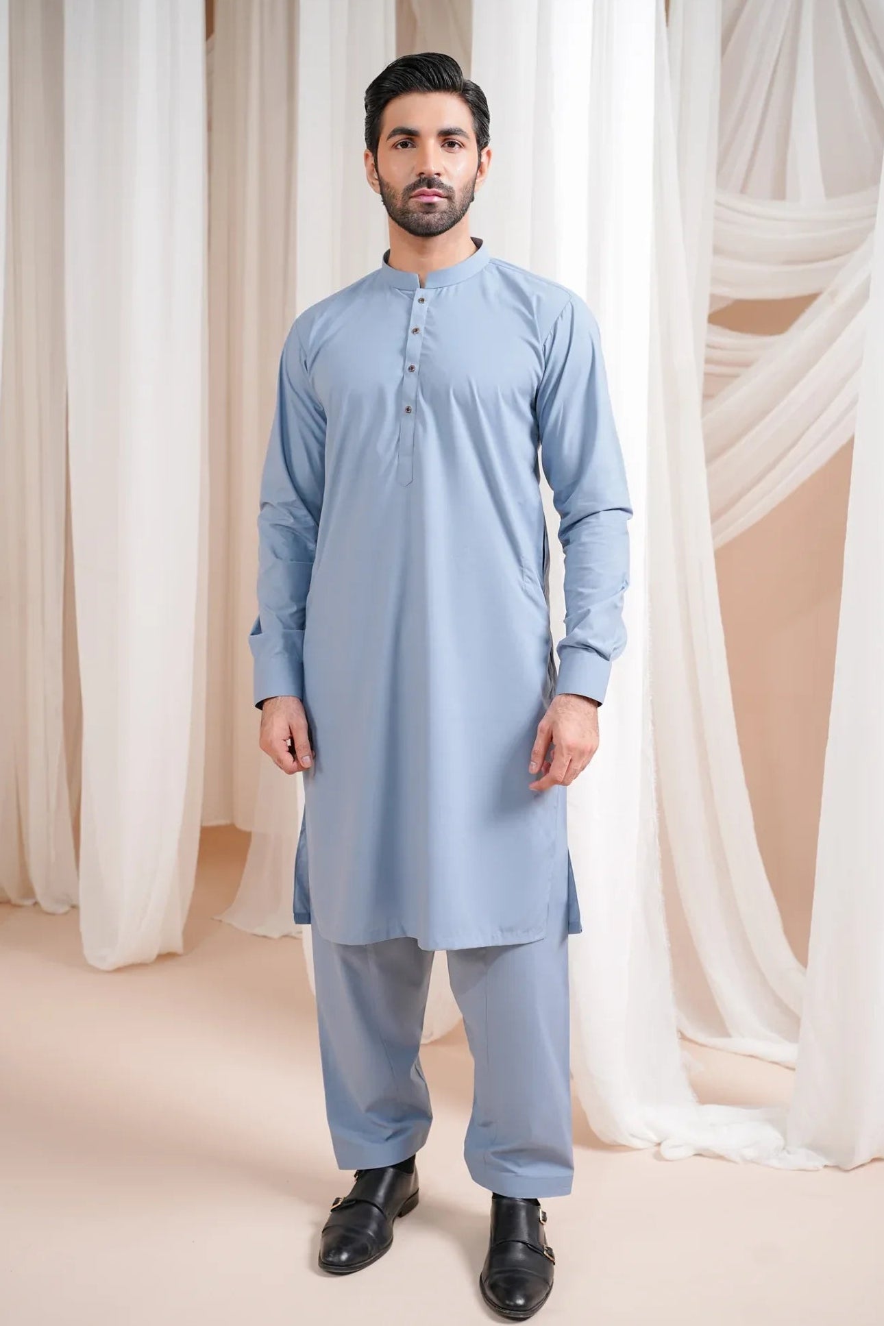 A man wearing a blue luxury blended kurta shalwar, standing against a curtain backdrop.