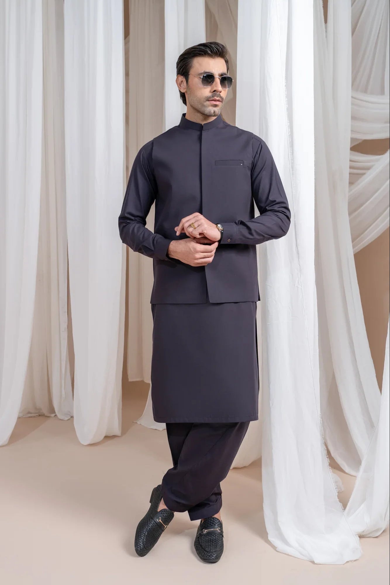 A man wearing a burgundy luxury formal kameez shalwar with a blended waistcoat and full sleeves. He is standing in a room with light curtains behind him.