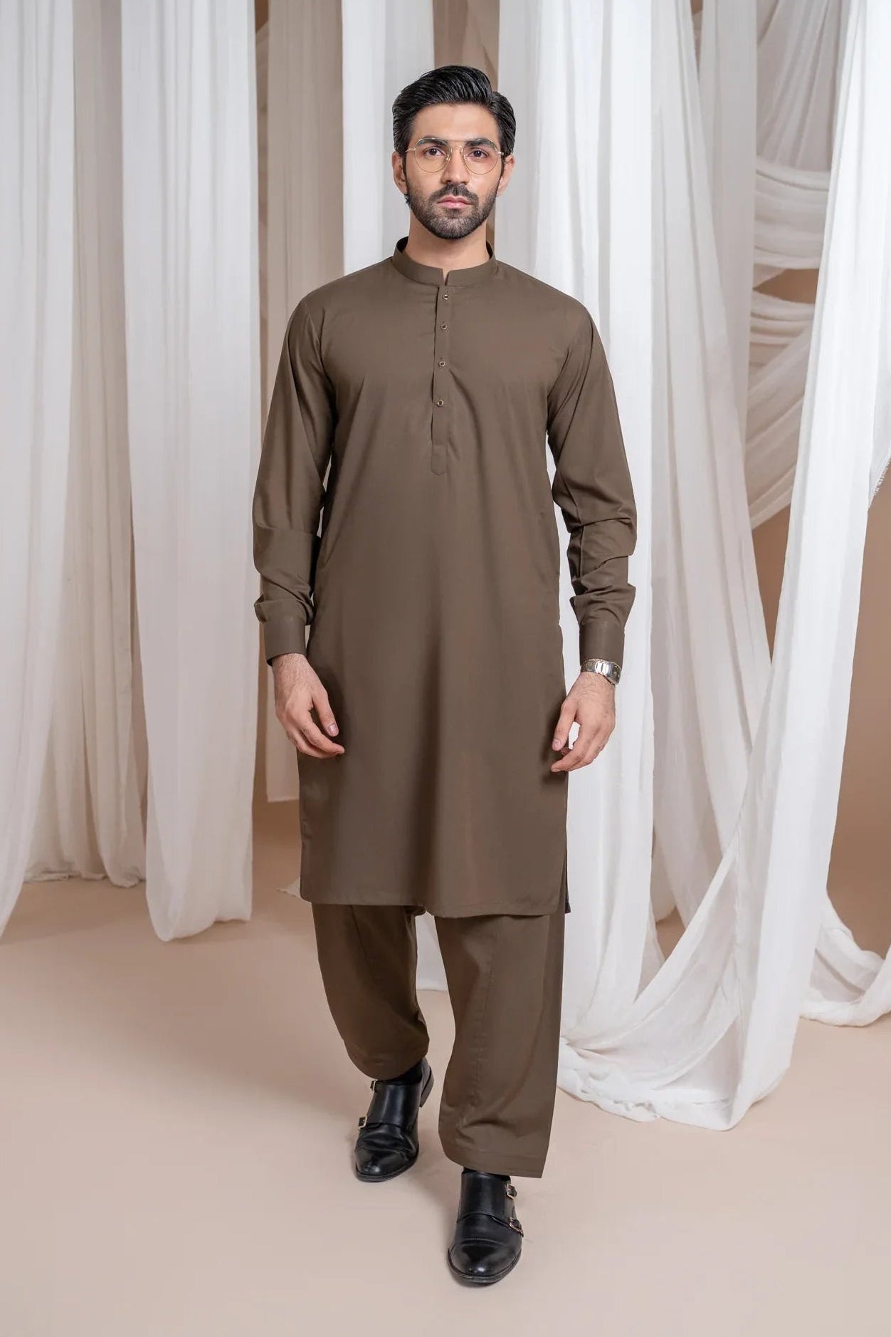 A man wearing a light brown luxury blended kurta shalwar set with a mandarin collar, full sleeves, and a straight kurta and trouser design, standing in a room with white draped backgrounds.