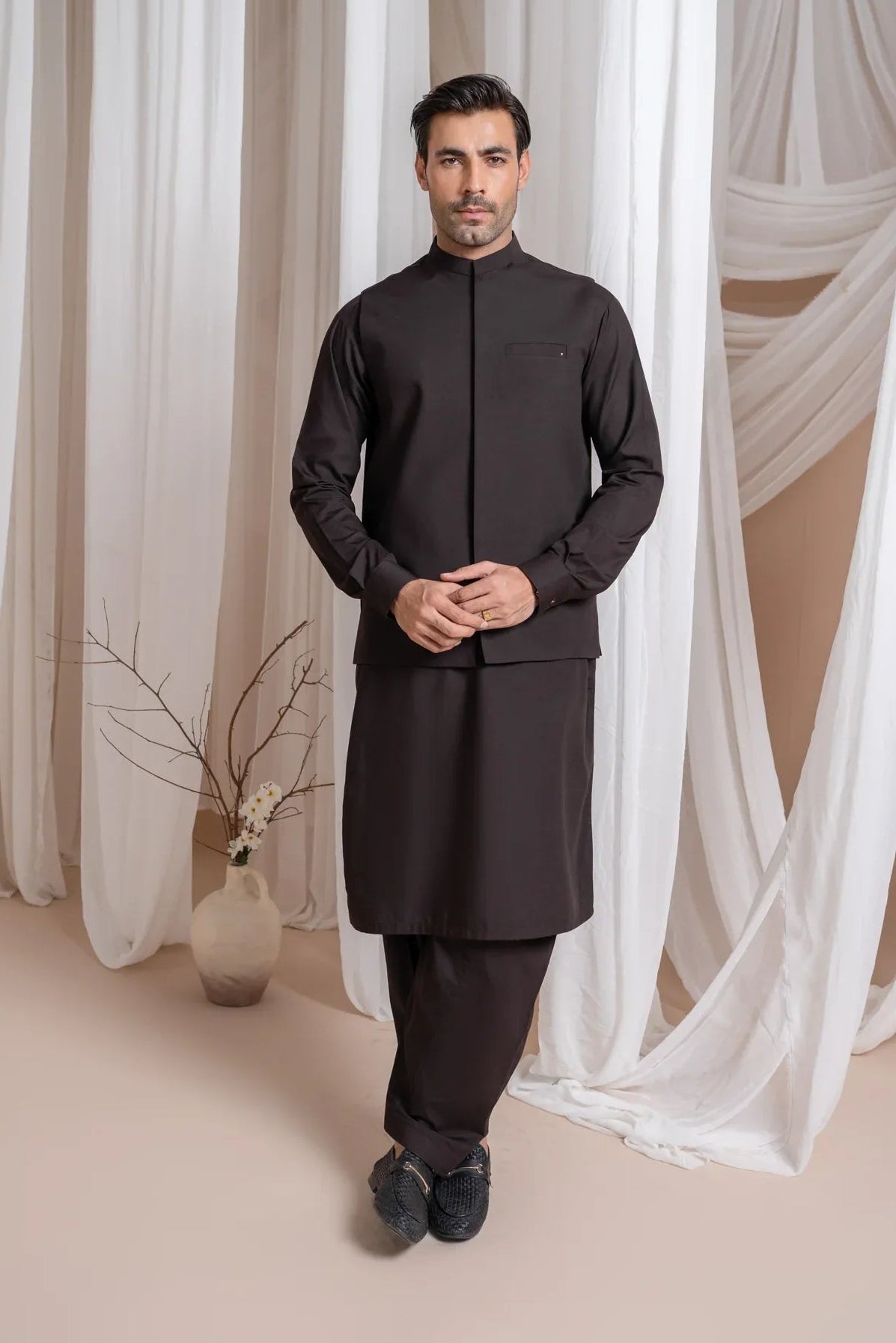 A man wearing a dark brown straight cut kameez shalwar with a blended waistcoat standing indoors with curtains in the background.