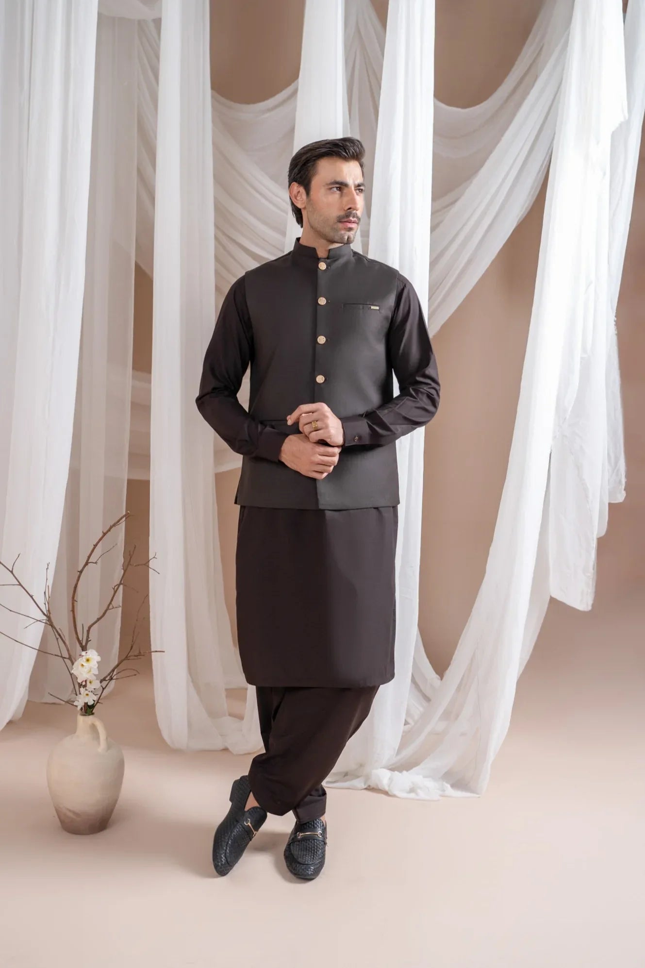 Man wearing a dark brown shalwar kameez and waistcoat with white curtains in the background