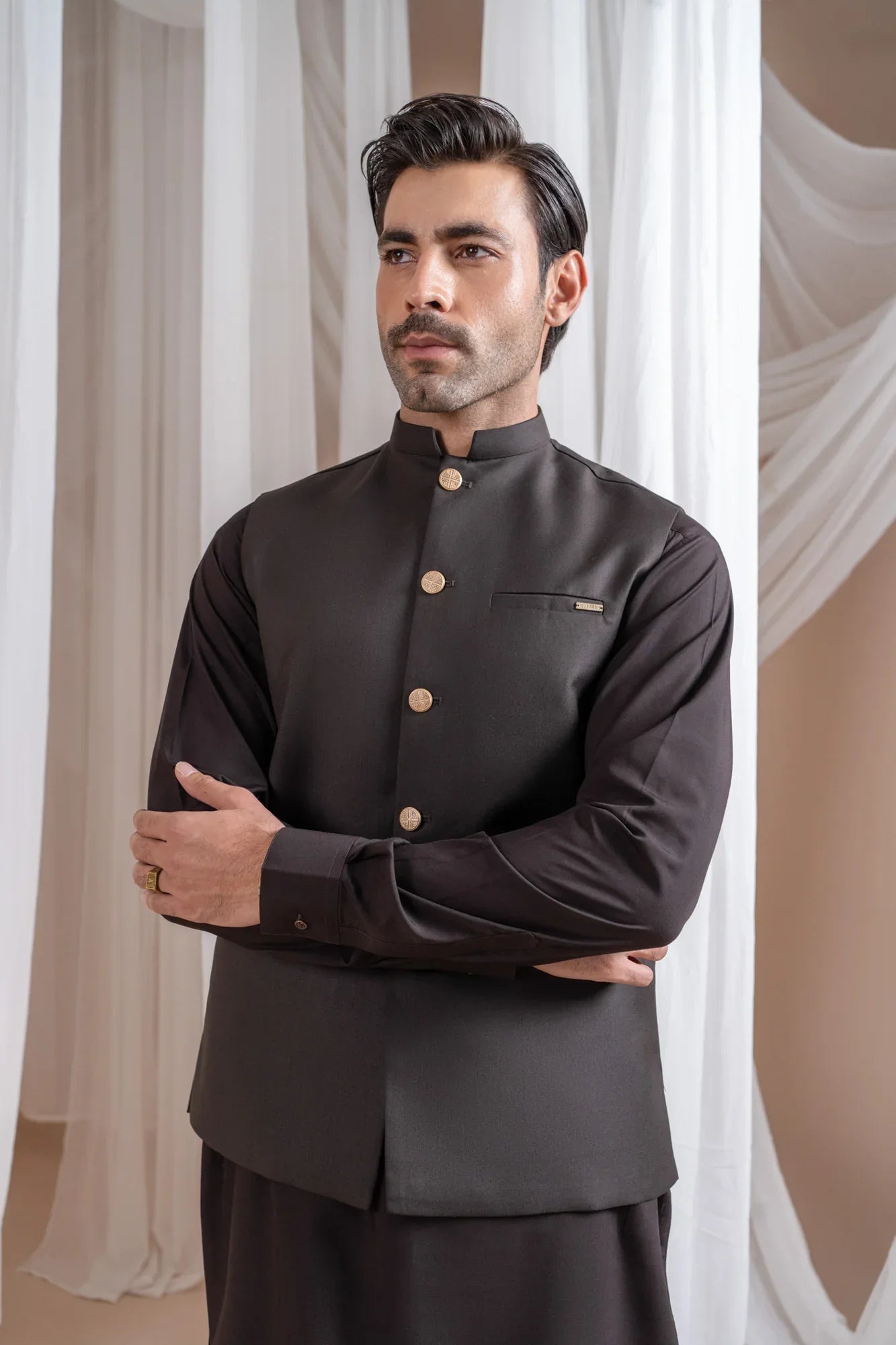 Man wearing a dark brown shalwar kameez and waistcoat with white curtains in the background