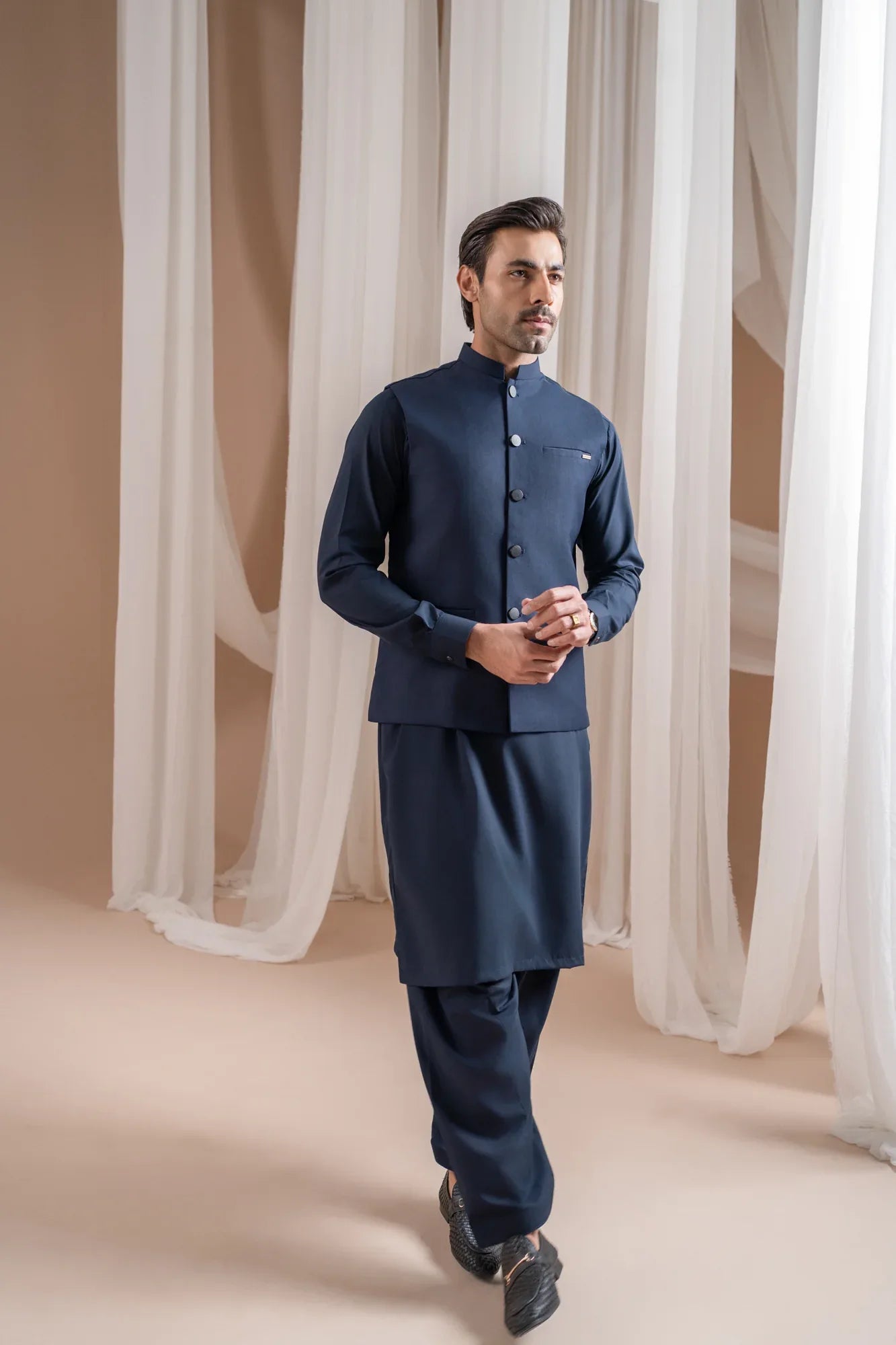 Man wearing a navy blue shalwar kameez with navy blue suiting waistcoat against a white curtain background