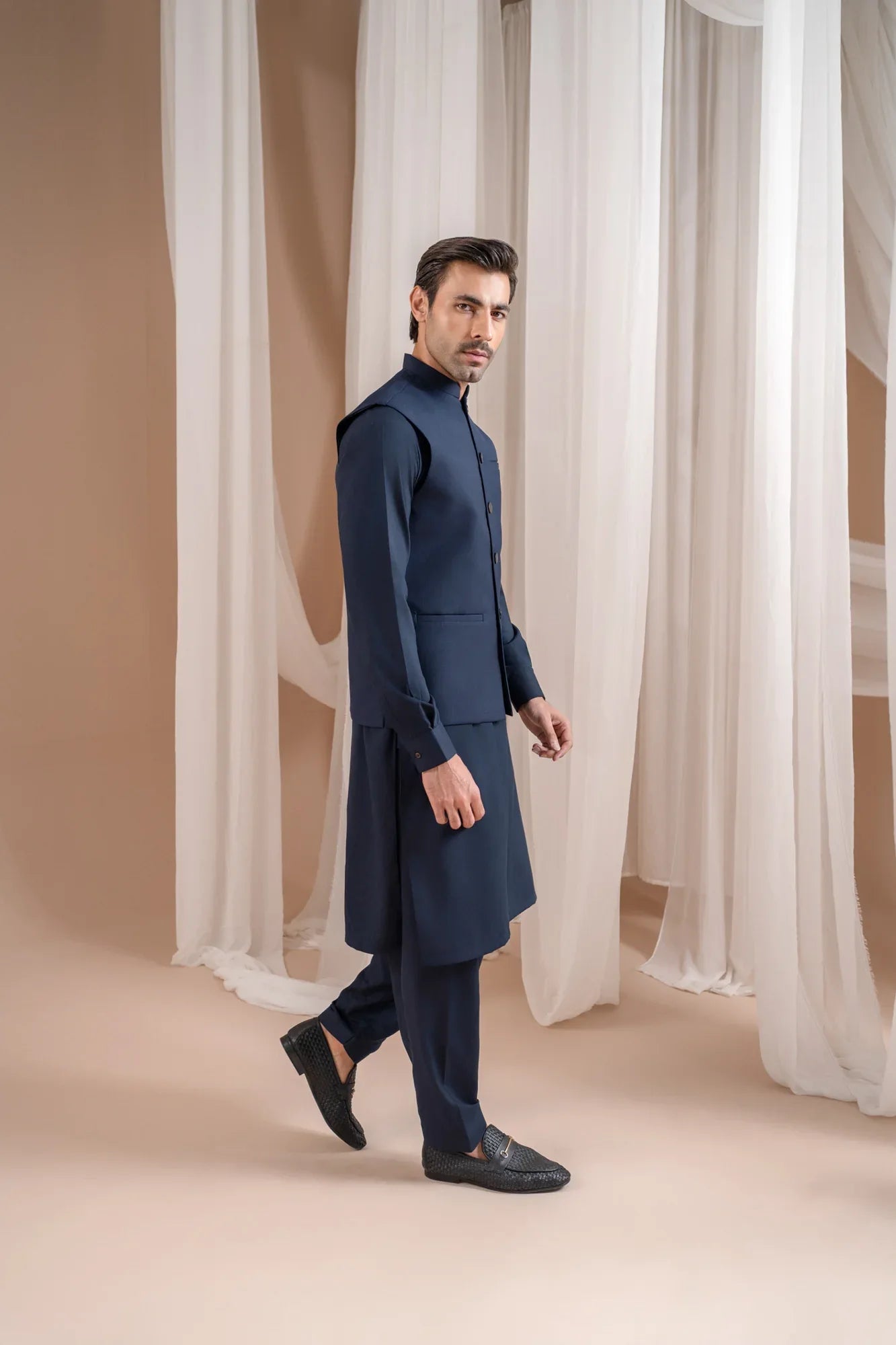 Man wearing a navy blue shalwar kameez with navy blue suiting waistcoat against a white curtain background