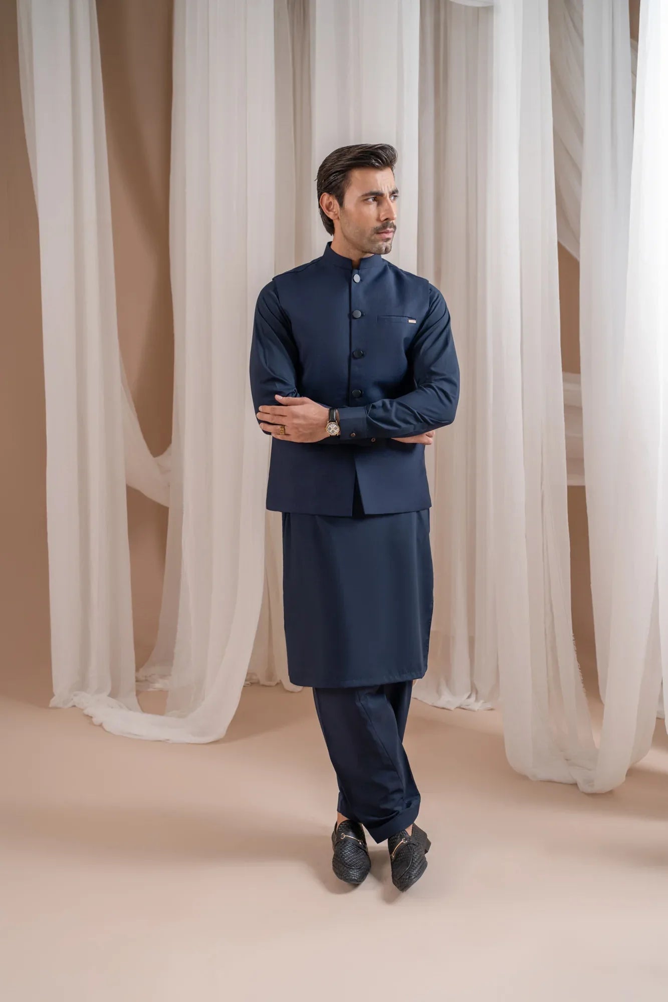 Man wearing a navy blue shalwar kameez with navy blue suiting waistcoat against a white curtain background