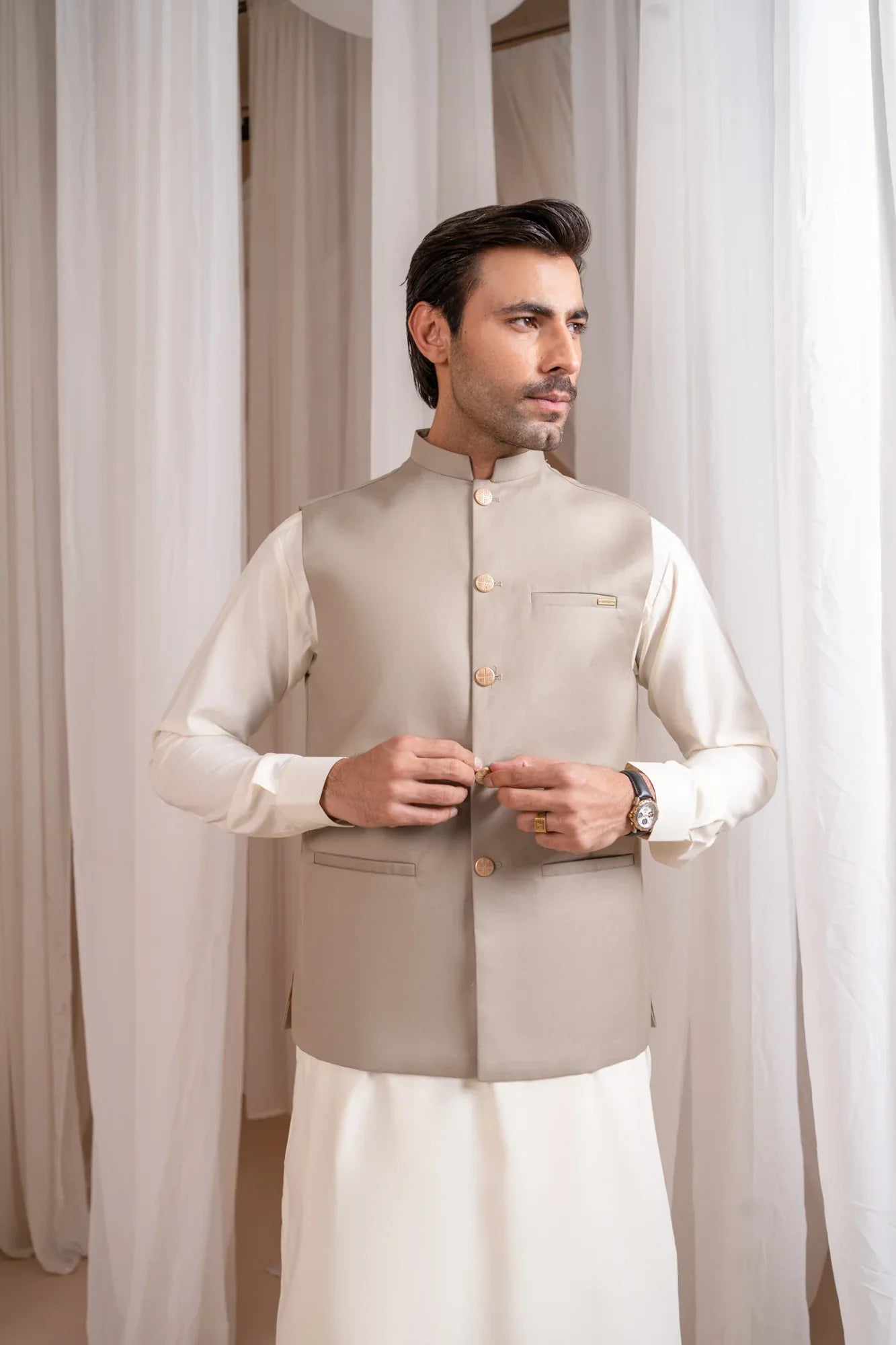 Man wearing a beige suiting waistcoat and white shalwar kameez against a white curtain background