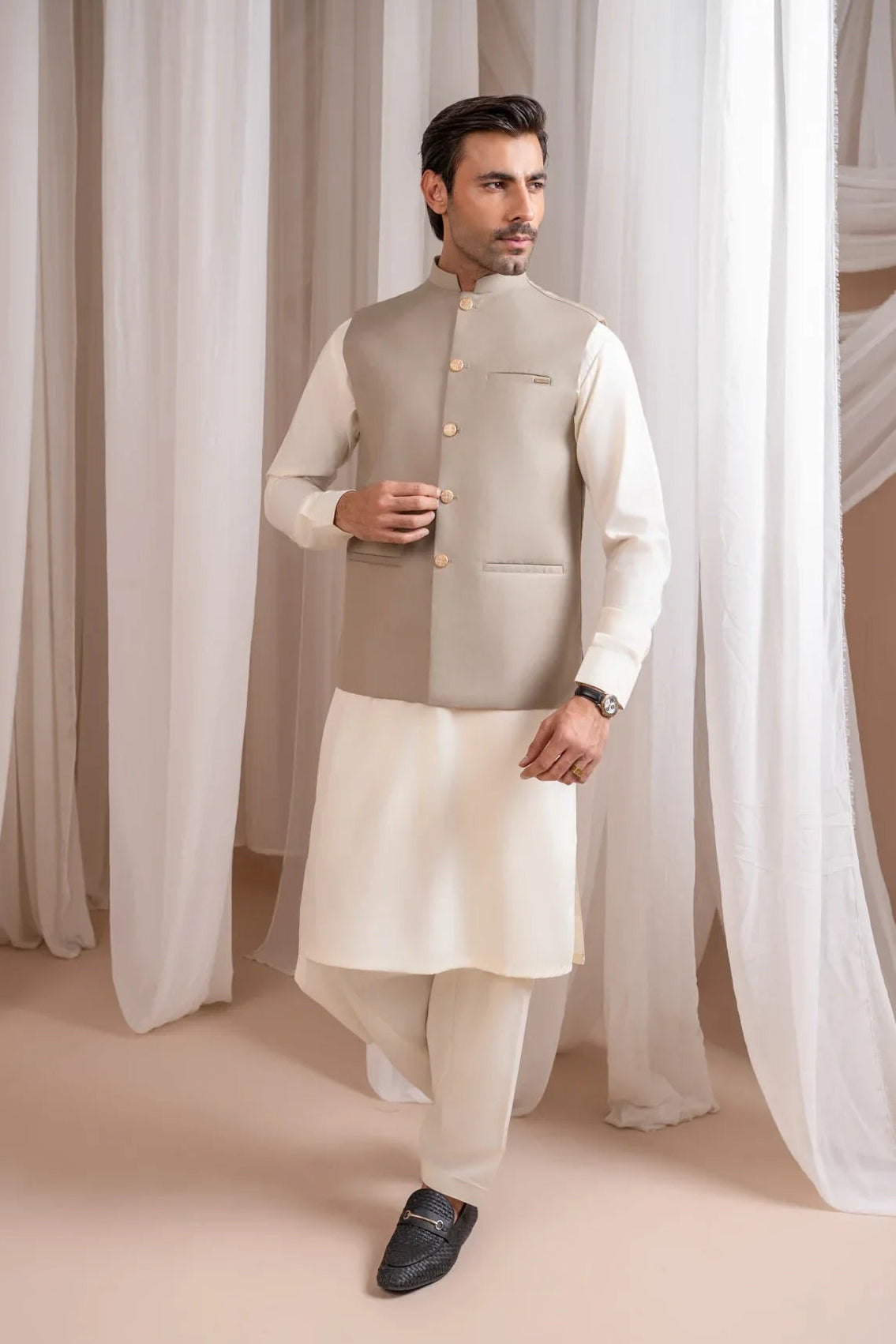 Man wearing a beige suiting waistcoat and white shalwar kameez against a white curtain background