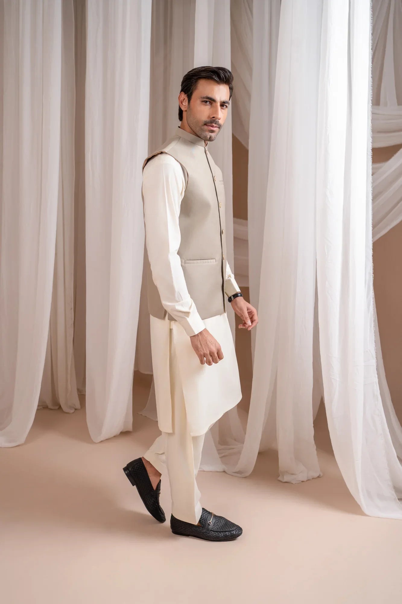 Man wearing a beige suiting waistcoat and white shalwar kameez against a white curtain background