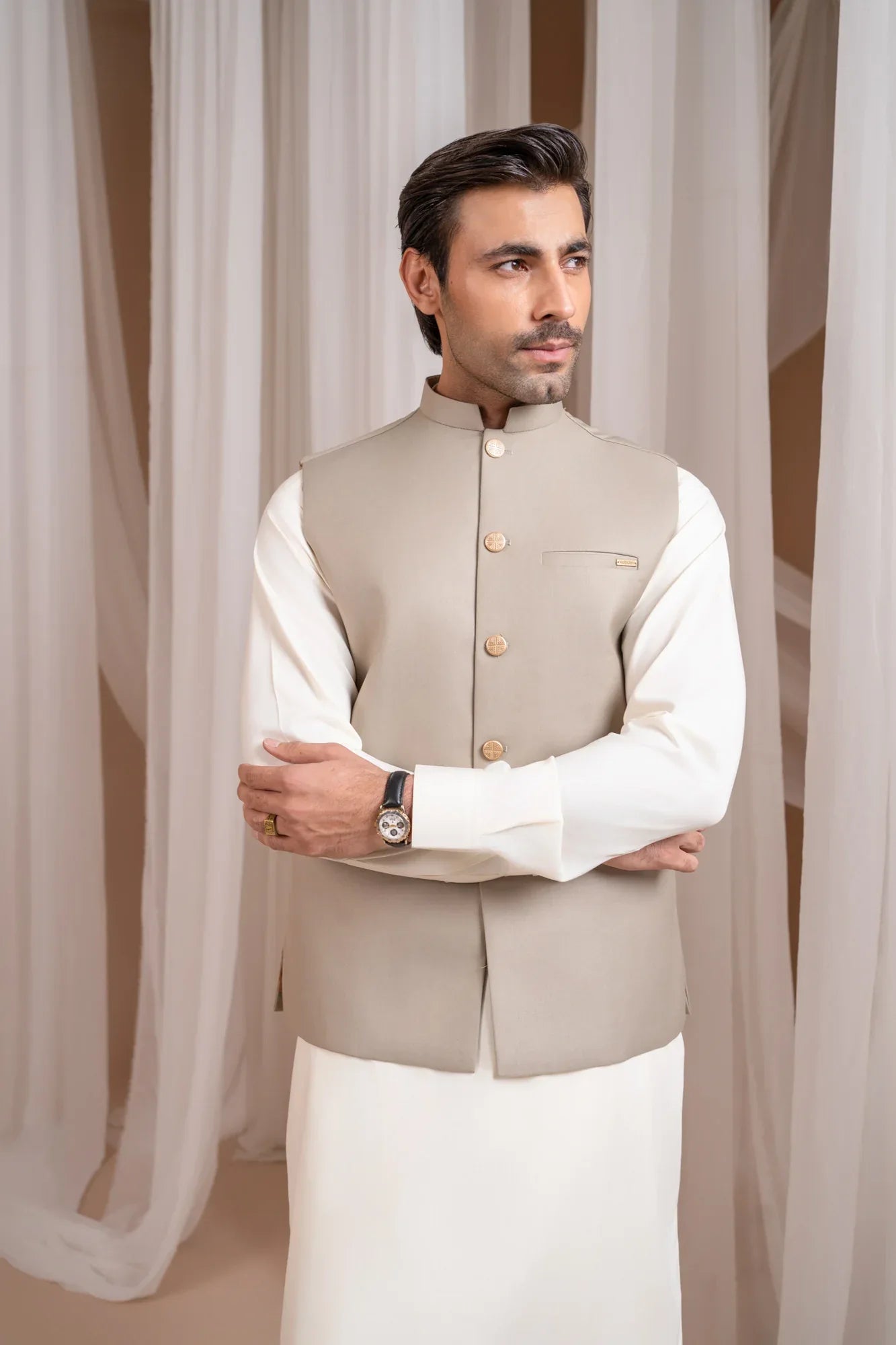 Man wearing a beige suiting waistcoat and white shalwar kameez against a white curtain background
