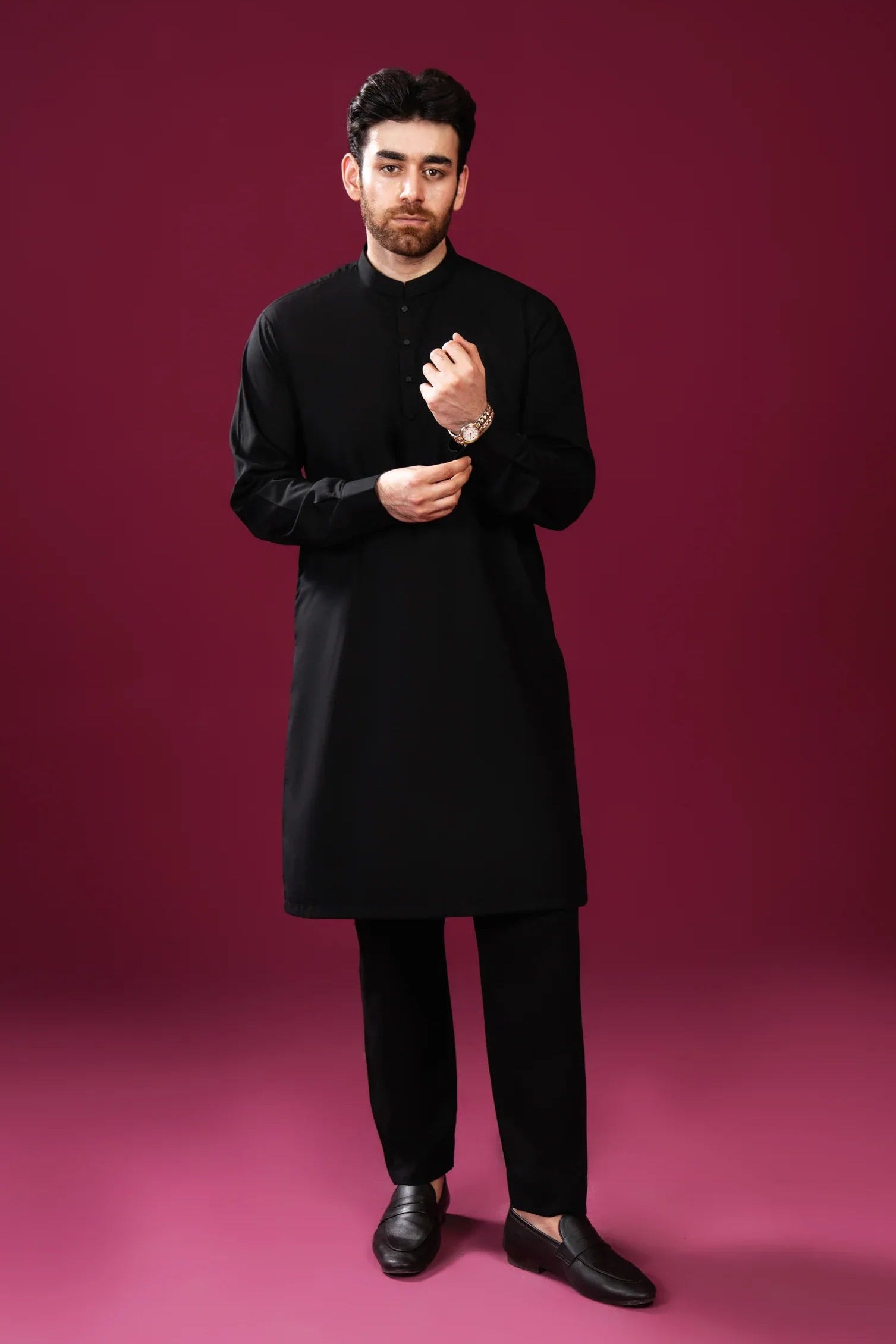 A man wearing a black kurta trouser with full sleeves and a mandarin collar, paired with black trouser/shalwar, standing against a pink background.