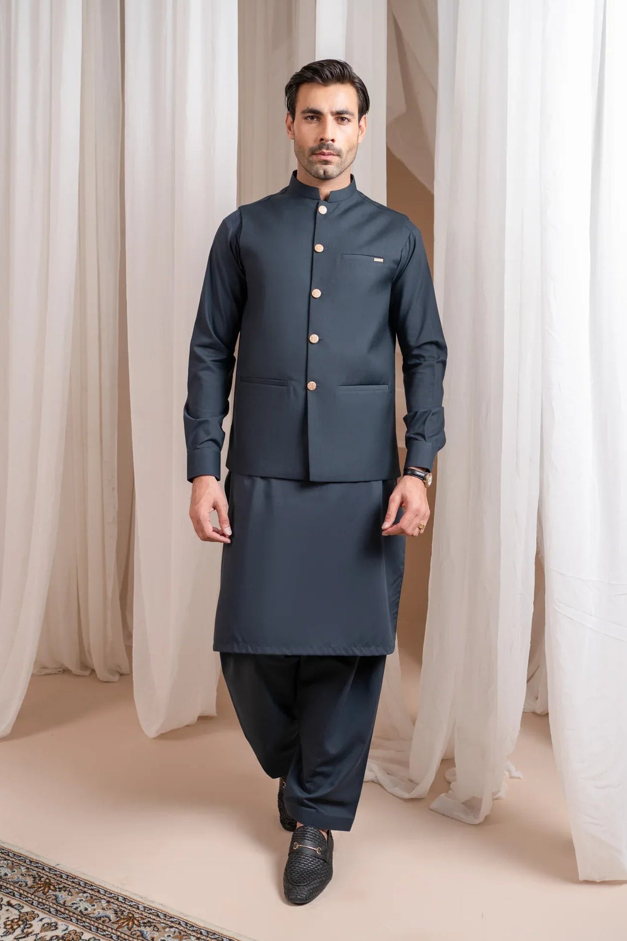 A man standing in a traditional attire, consisting of a charcoal grey kurta shalwar and a matching suiting waistcoat, against a backdrop with white drapes.