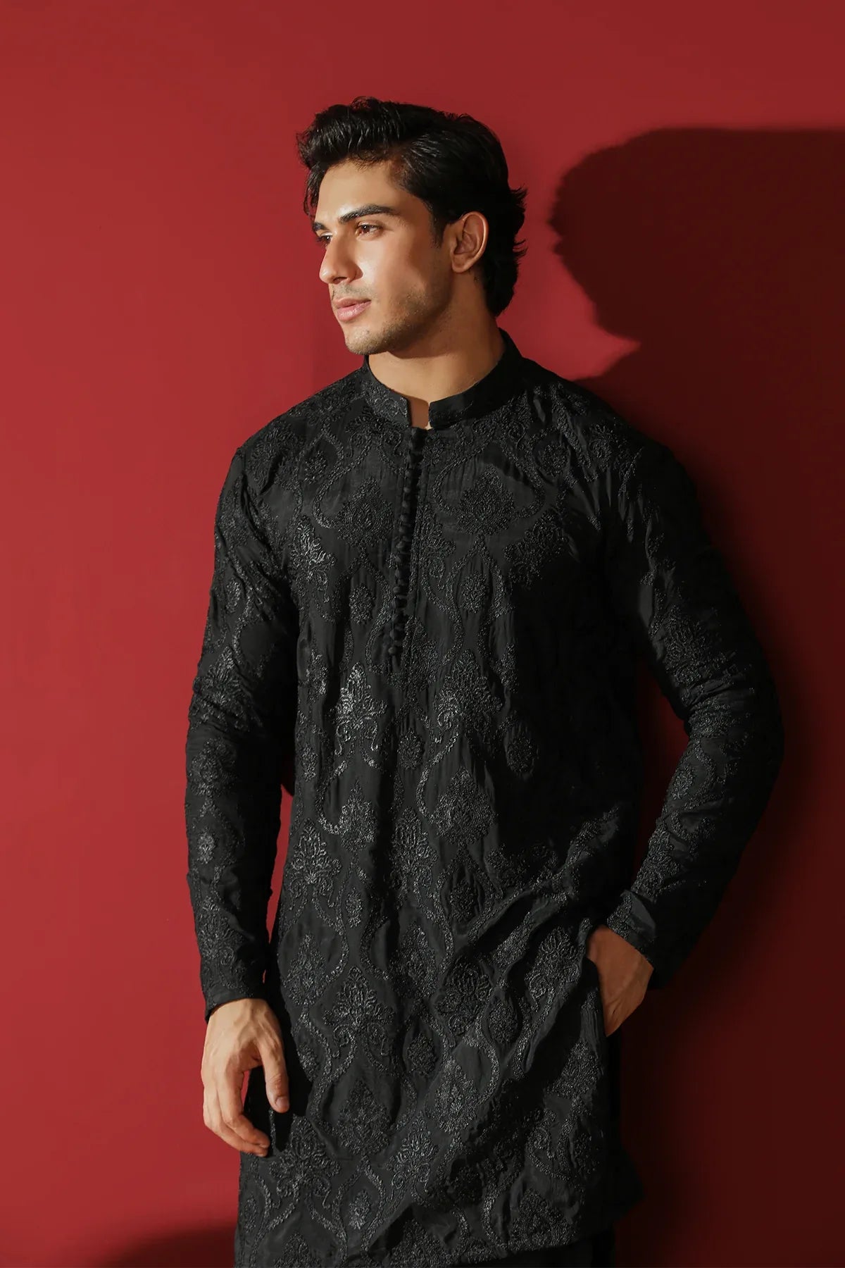 Man wearing a black raw silk embroidered kurta trouser against a red background